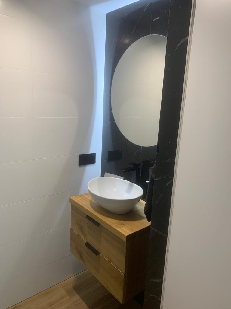Nowoczesna łazienka z umywalką nablatową na drewnianej szafce z czarnymi uchwytami, okrągłe lustro oprawione w czarny marmur, białe płytki na ścianie, drewniana podłoga