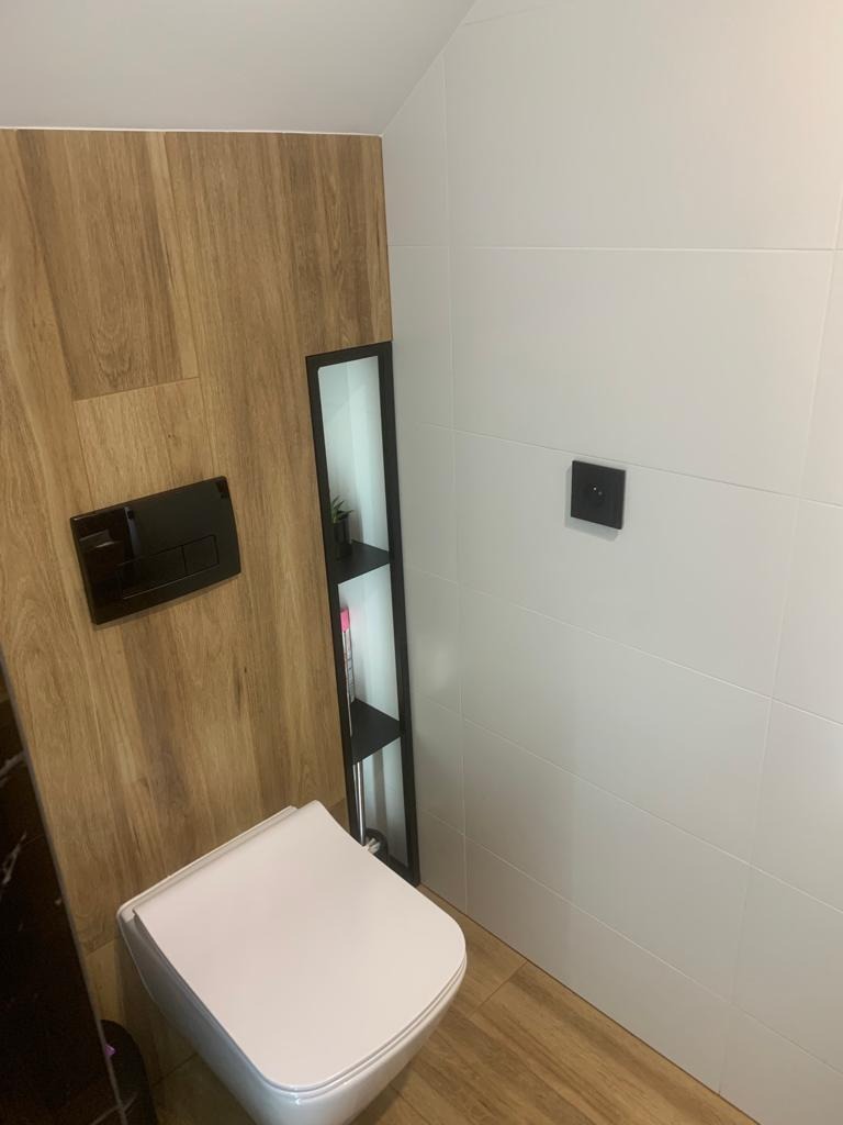 Nowoczesna toaleta podwieszana z drewnopodobną obudową, wbudowana półka z czarną ramą, białe płytki na ścianie i podłodze.