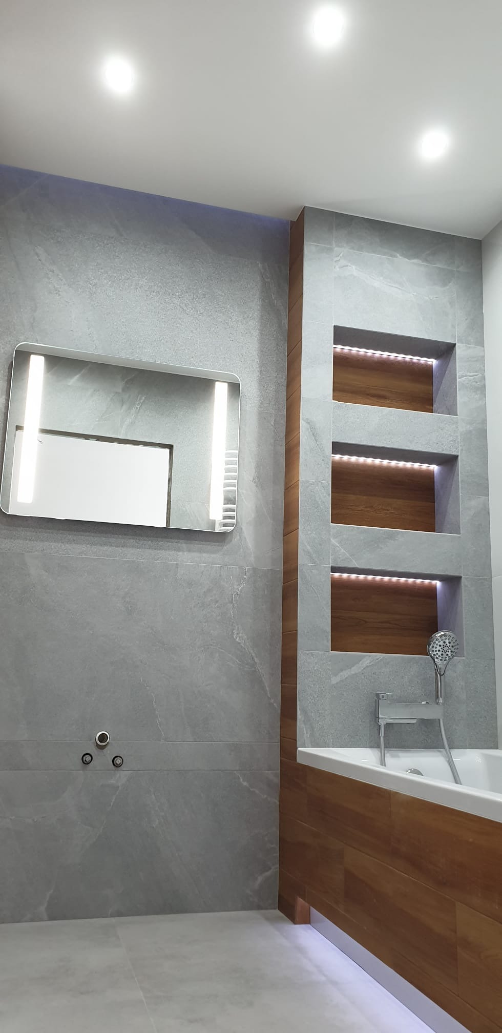 Nowoczesna łazienka z szarymi płytkami imitującymi kamień, podświetlanym lustrem, drewnianymi wnękami z oświetleniem LED i wanną obudowaną drewnem.