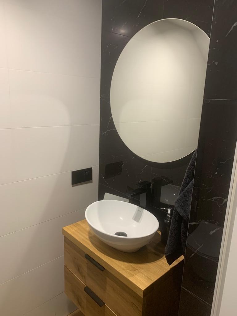 Nowoczesna łazienka z białą umywalką nablatową na drewnianej szafce, czarną armaturą i okrągłym lustrem wkomponowanym w czarne płytki imitujące marmur, kontrastujące z białymi płytkami na ścianie.