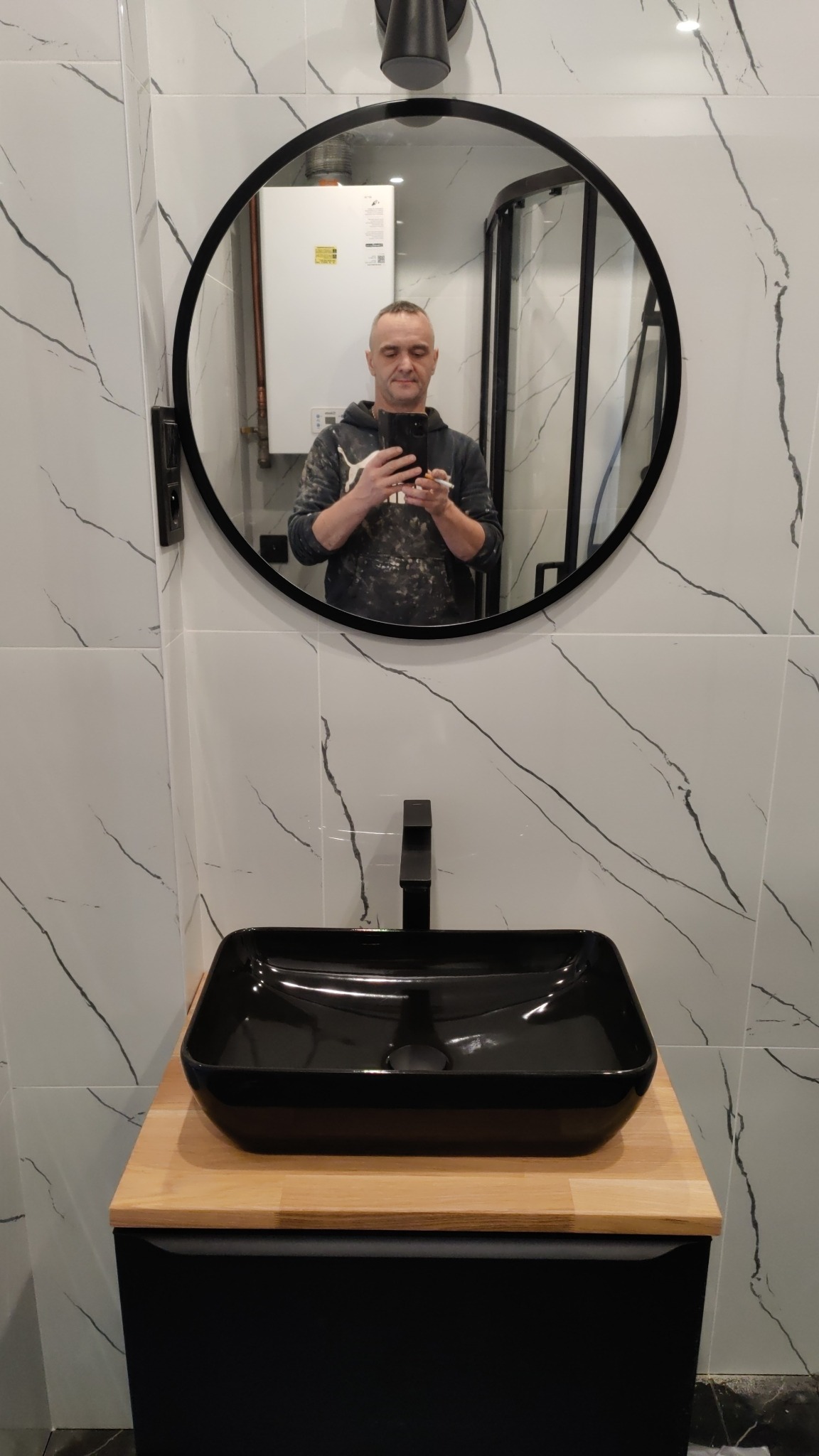 Nowoczesna łazienka w Katowicach z czarną umywalką nablatową i baterią, marmurowe płytki na ścianach, okrągłe lustro z odbiciem osoby robiącej zdjęcie.