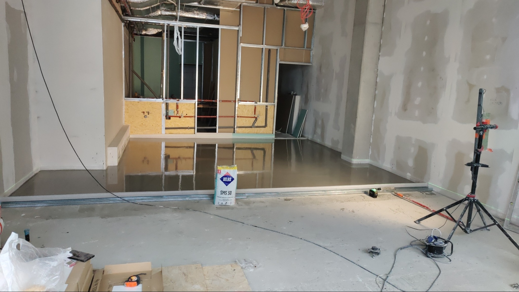 Wylewka samopoziomująca na podłodze w trakcie prac wykończeniowych, widoczny worek z zaprawą Atlas SMS 30, metalowe profile, laser krzyżowy i lampa na statywie.