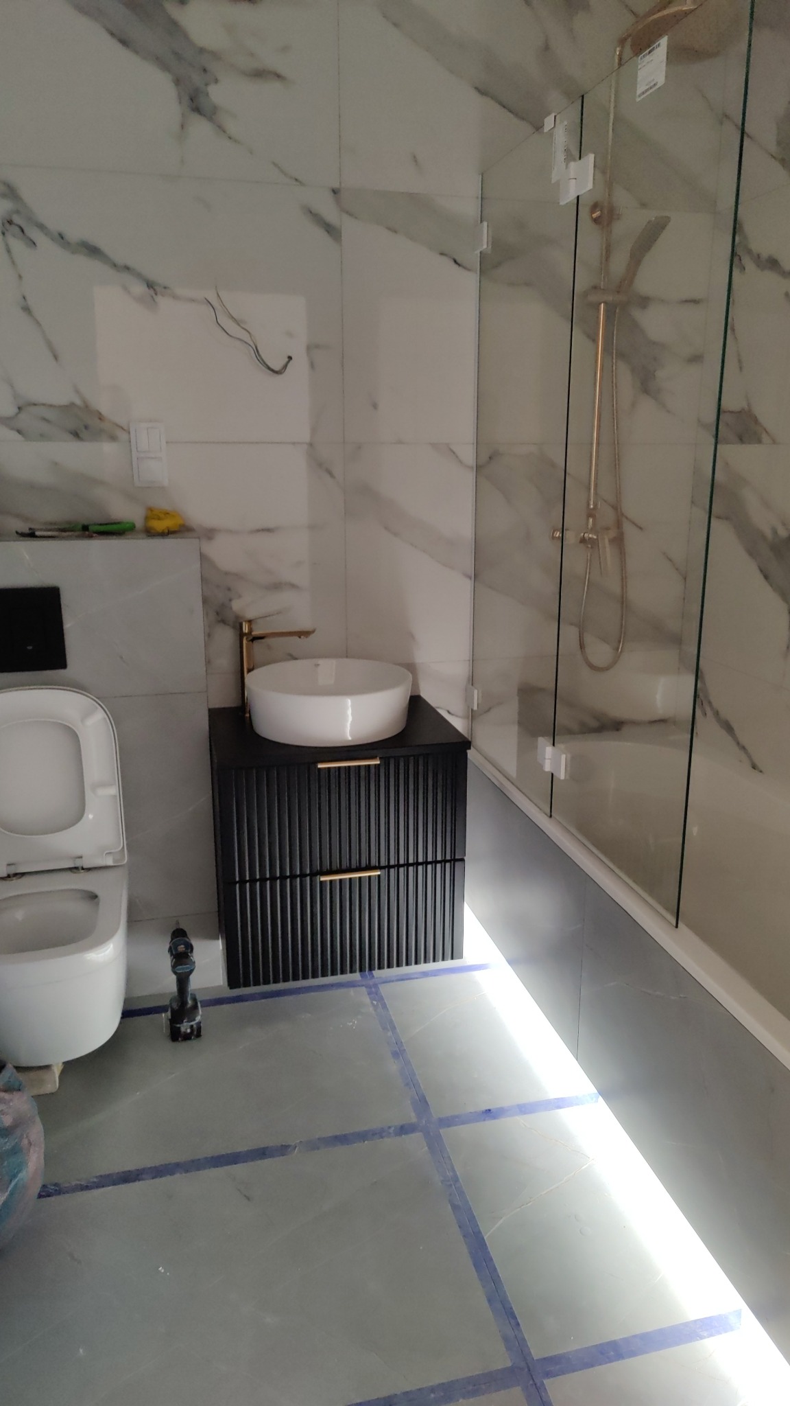 Nowoczesna łazienka w trakcie wykańczania: marmurowe płytki ścienne, czarna szafka pod umywalką z frezowanymi frontami i złotymi uchwytami, okrągła biała umywalka nablatowa, kabina prysznicowa ze...