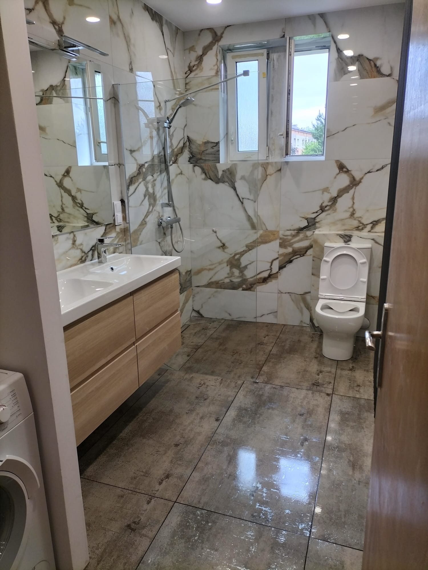 Wykończona łazienka z marmurowymi płytkami na ścianach, drewnianą szafką pod umywalką, toaletą i szarą podłogą z płytek, widok na okno z widokiem na zewnątrz.