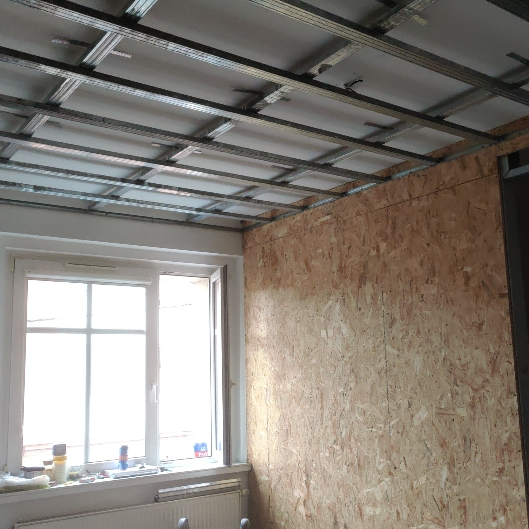 Wnętrze pokoju w trakcie prac wykończeniowych: metalowy stelaż sufitu podwieszanego zamontowany, ściana wyłożona płytą OSB, okno z widocznymi przedmiotami na parapecie.