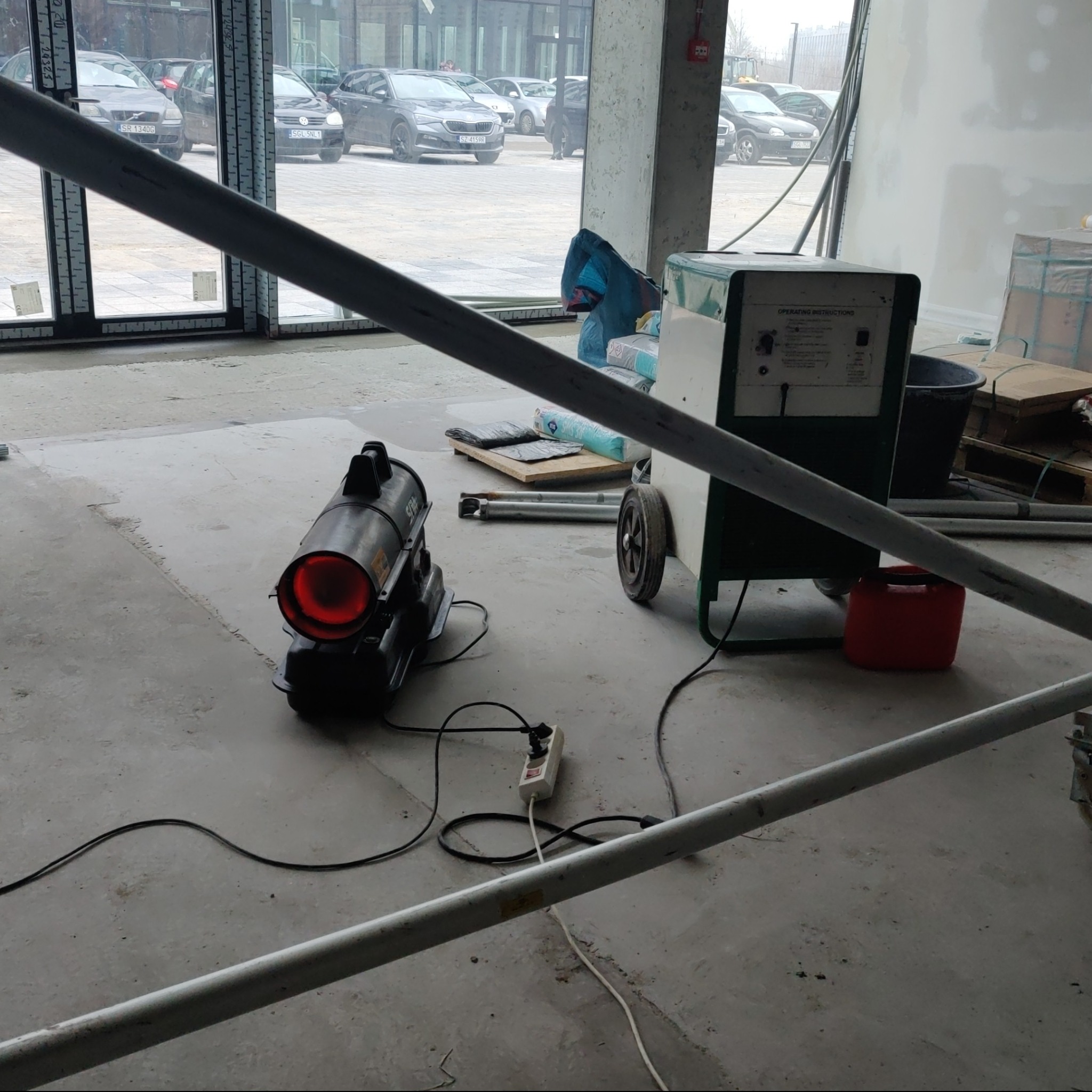 Wnętrze w trakcie prac wykończeniowych, widoczny nagrzewnica elektryczna, agregat malarski na kółkach, worki z materiałami budowlanymi oraz fragment rusztowania na pierwszym planie.