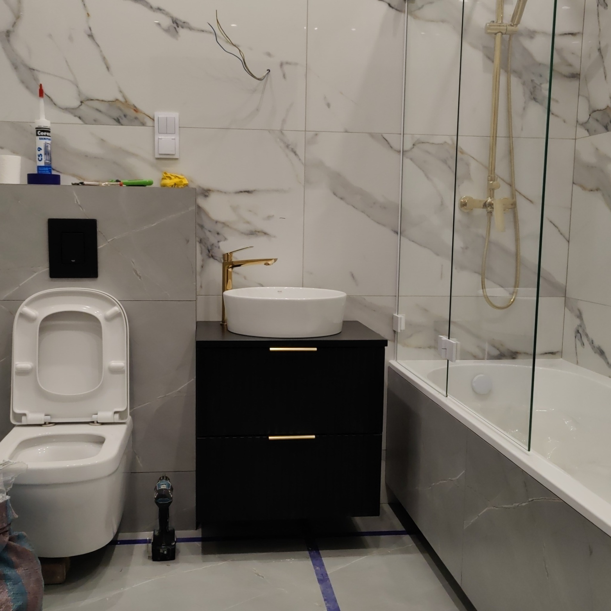 Nowoczesna łazienka w trakcie wykańczania: biała umywalka na czarnej szafce z złotą armaturą, toaleta z otwartą klapą, wanna za szklanym parawanem, marmurowe płytki na ścianach, widoczne narzędzia...