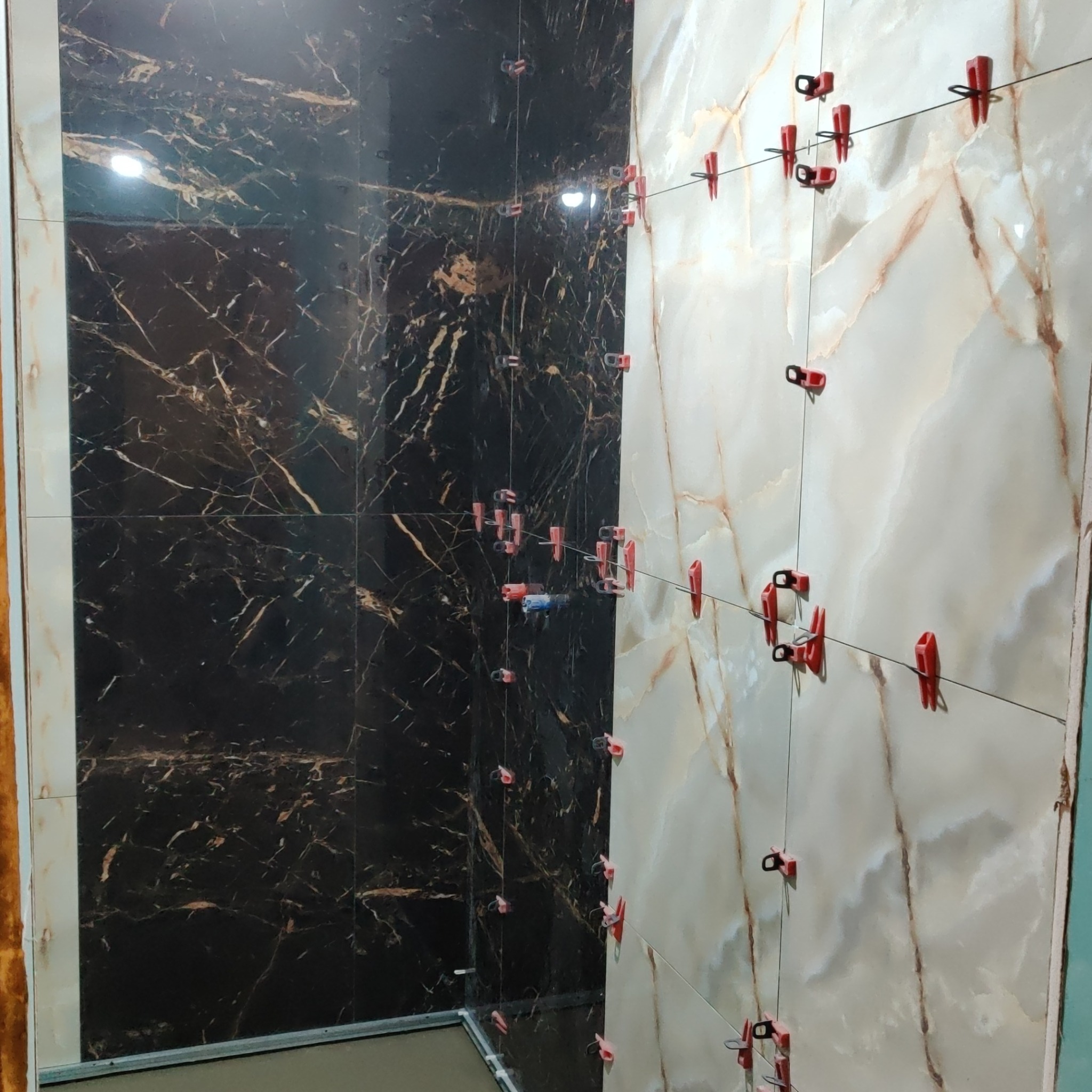 Proces układania płytek w łazience, widoczne płytki imitujące marmur w dwóch kolorach - ciemnym i jasnym, oraz system poziomowania płytek z czerwonymi klipsami i sznurkiem.