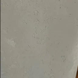 Szara ściana z wyraźną, nieregularną fakturą. Drobne wgłębienia i wypukłości tworzą unikalny wzór na powierzchni. Stonowana kolorystyka, bliski kadr.