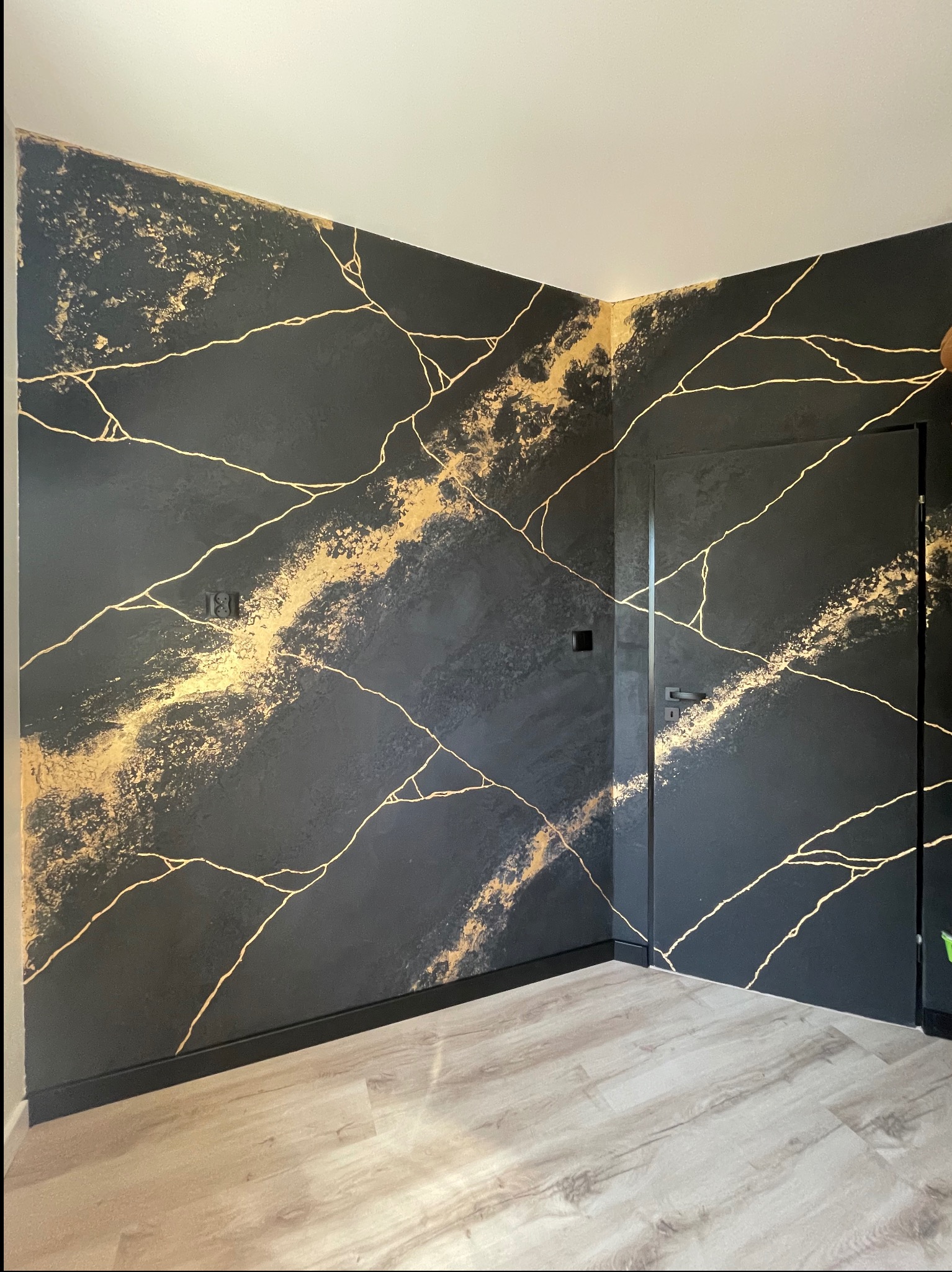 Ściana z efektem czarnego marmuru i złotych żył, widoczna w rogu pokoju. Dekoracyjne wykończenie wnętrza z dopasowanymi listwami przypodłogowymi i drzwiami.