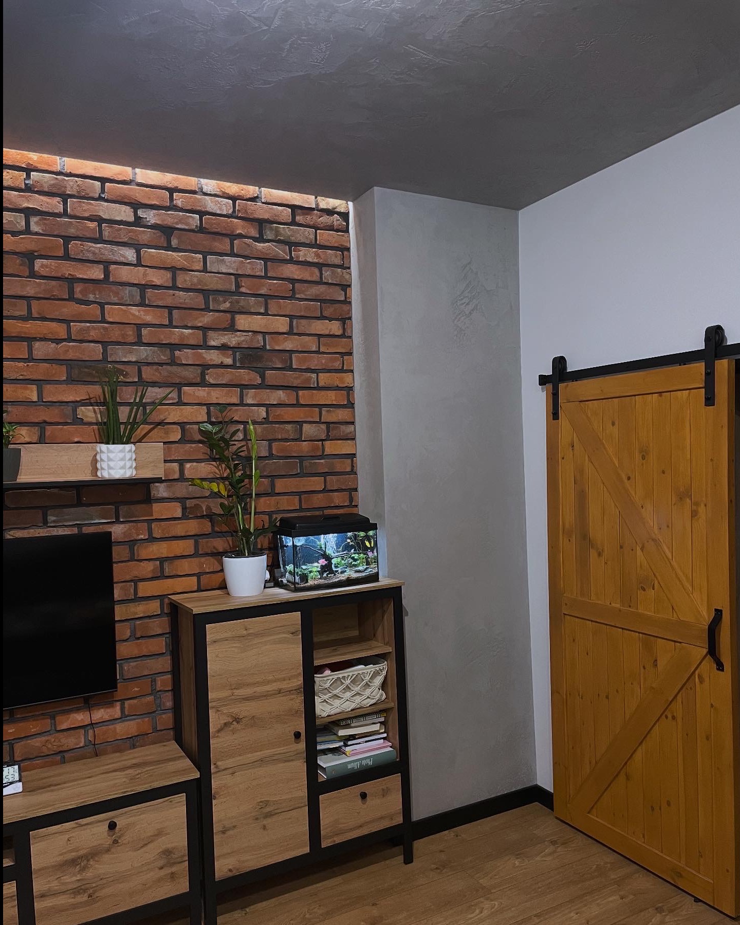 Loftowe wnętrze: ściana z czerwonej cegły, drewniane meble z czarnymi akcentami, akwarium i przesuwne drzwi w stylu rustykalnym. Dekoracyjna szara ściana.