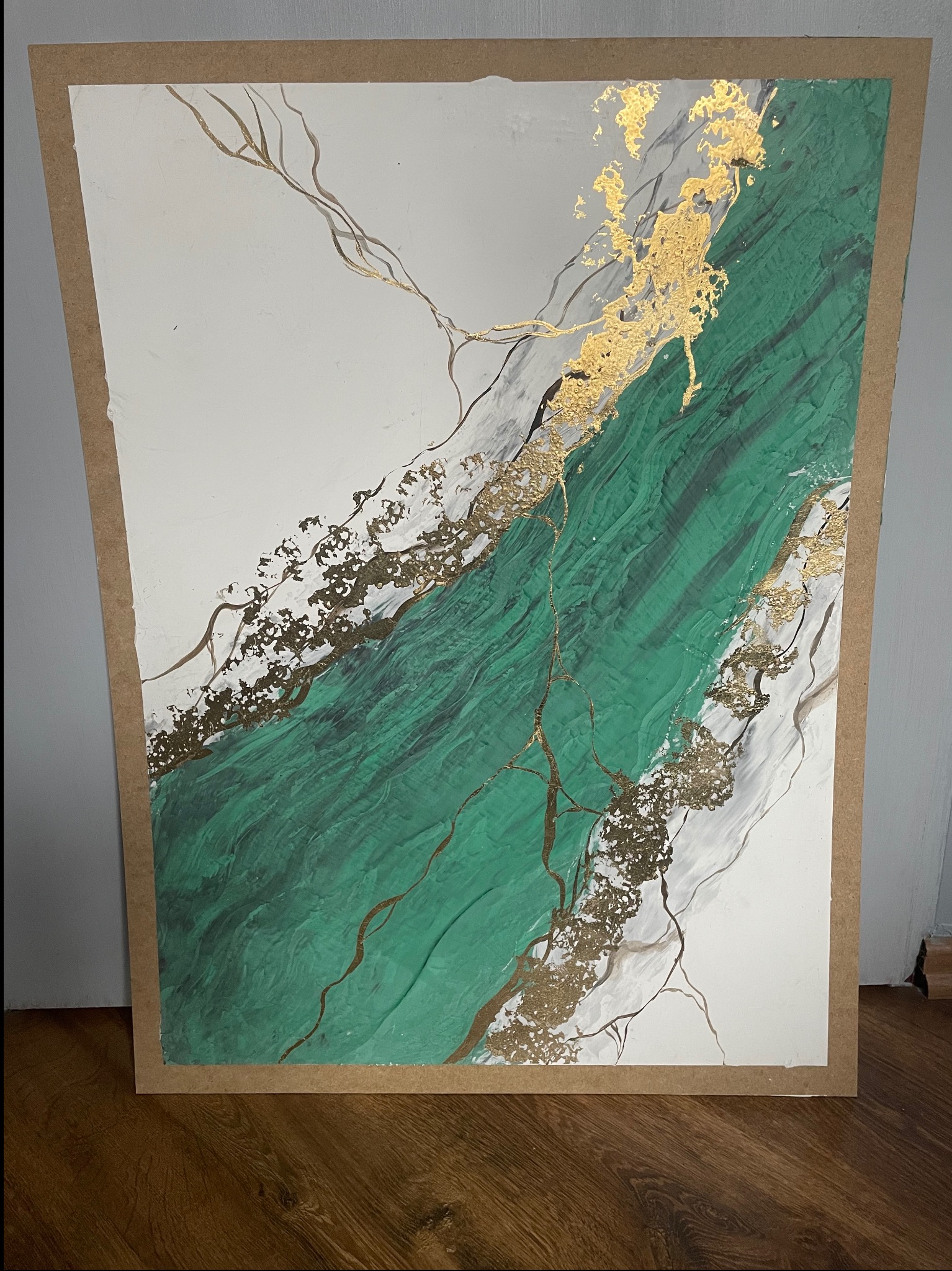 Abstrakcyjny obraz z zielonymi i złotymi akcentami, imitujący marmur, w brązowej ramie, na tle podłogi z paneli. Wyjątkowa faktura i połyskujące detale.
