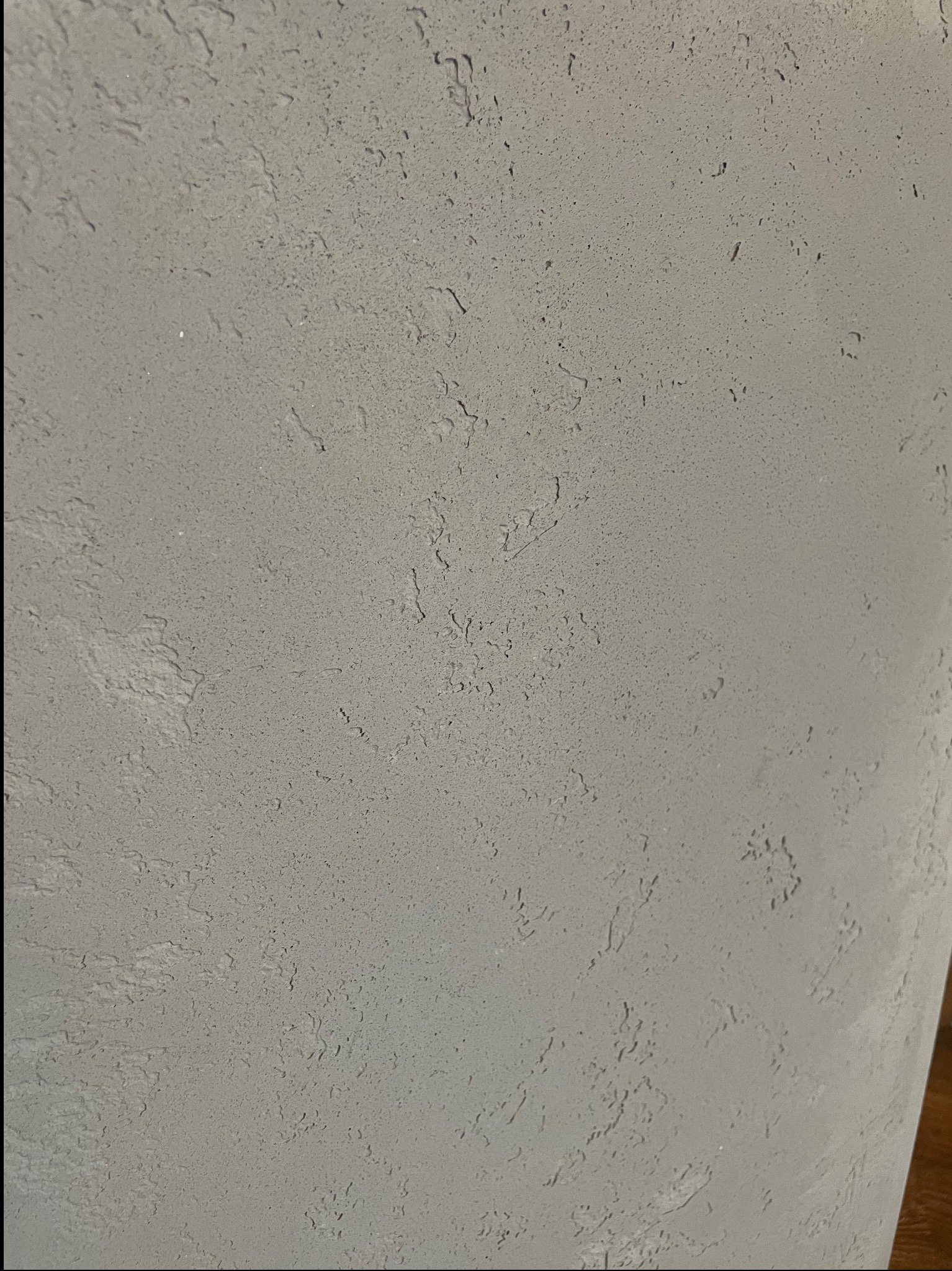 Szara ściana z wyraźną, nieregularną fakturą. Drobne wgłębienia i wypukłości tworzą unikalny wzór na powierzchni. Stonowana kolorystyka, bliski kadr.