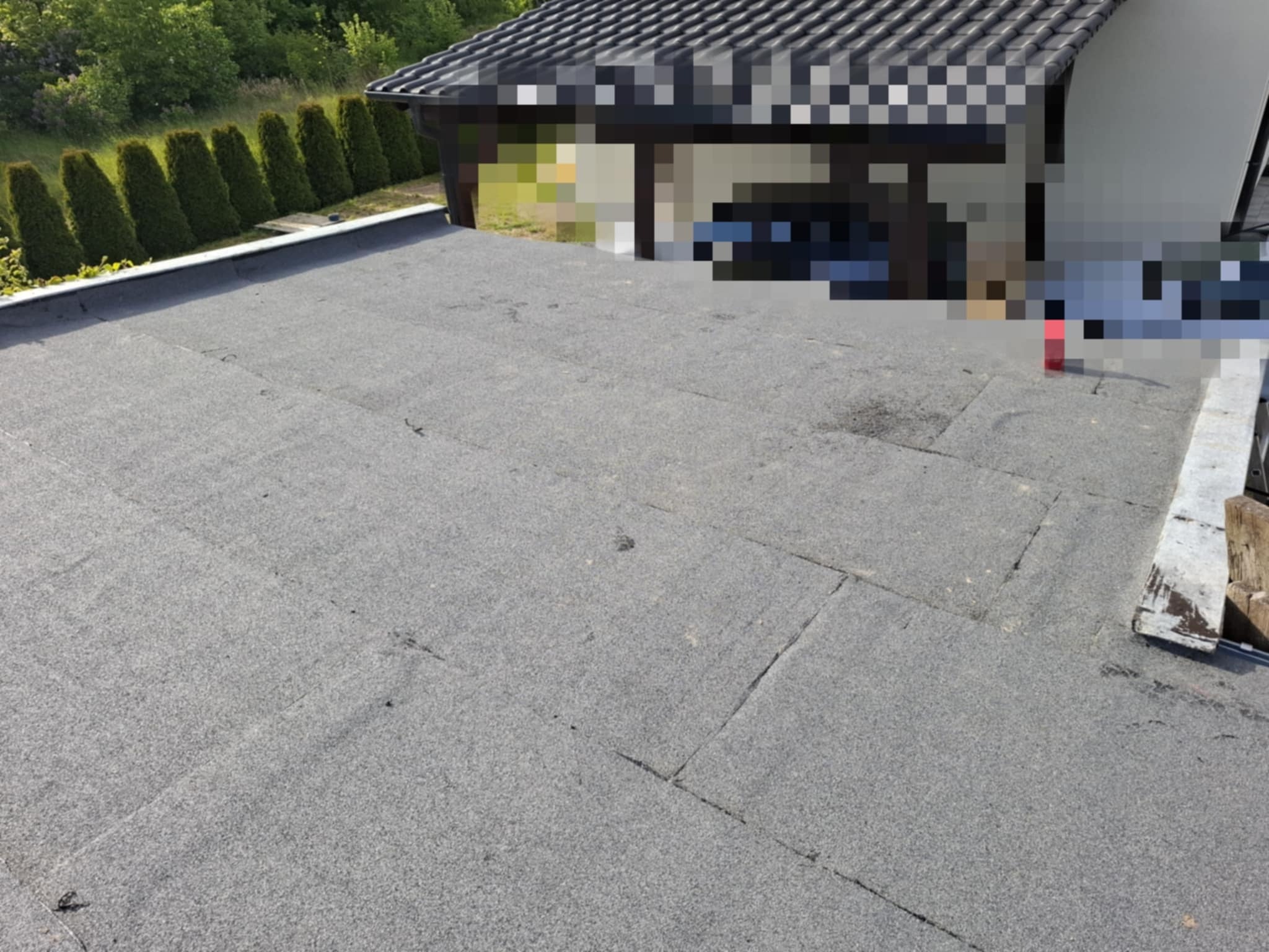 Szara papa na płaskim dachu domu, widok z góry, z fragmentem dachu garażu i rzędem tuj w tle. Widoczne łączenia papy i fragment obróbki blacharskiej.