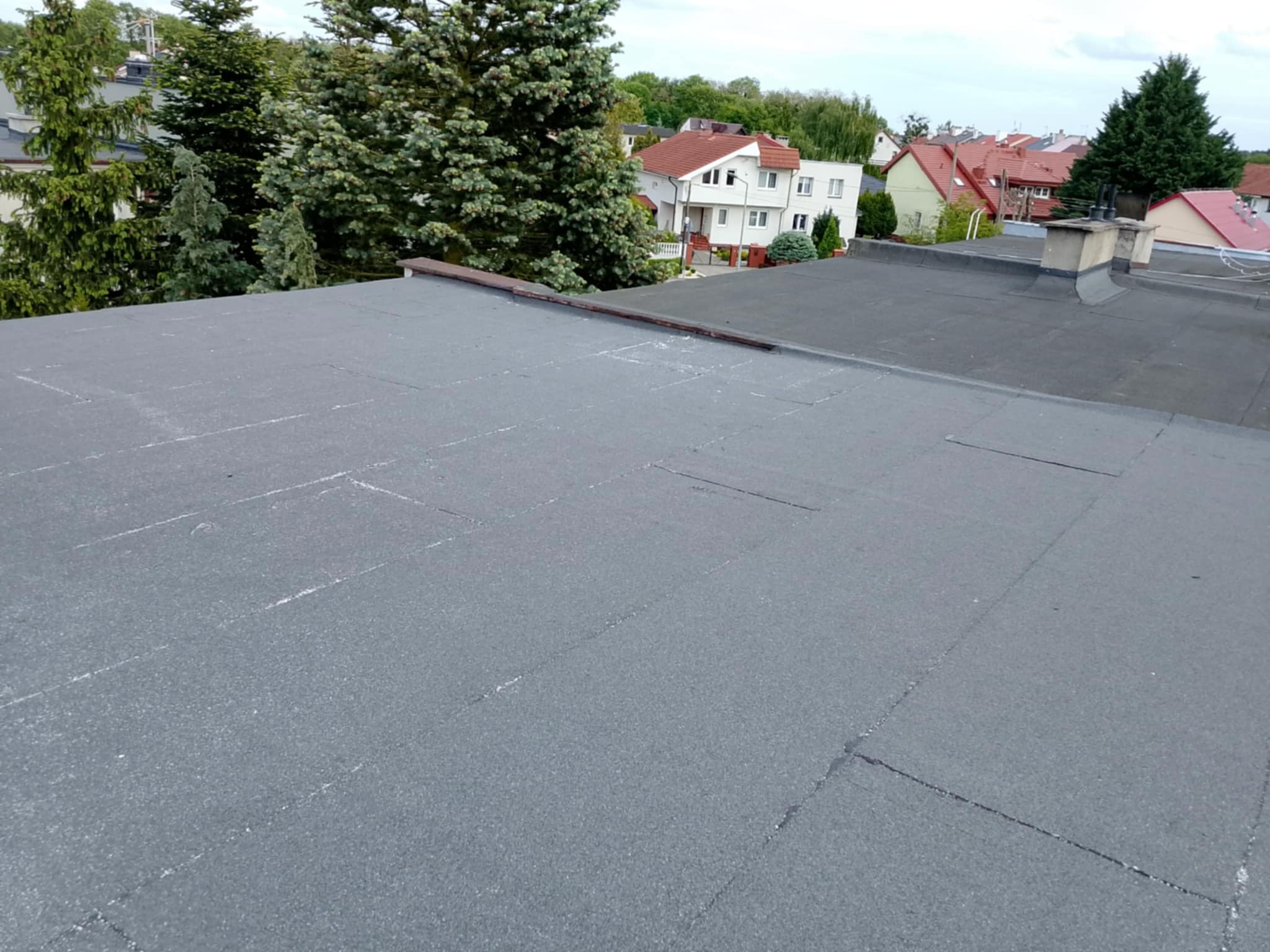 Szary, płaski dach pokryty papą na tle zieleni drzew i zabudowań. Widoczne łączenia papy. Ujęcie z góry, podkreślające strukturę pokrycia.