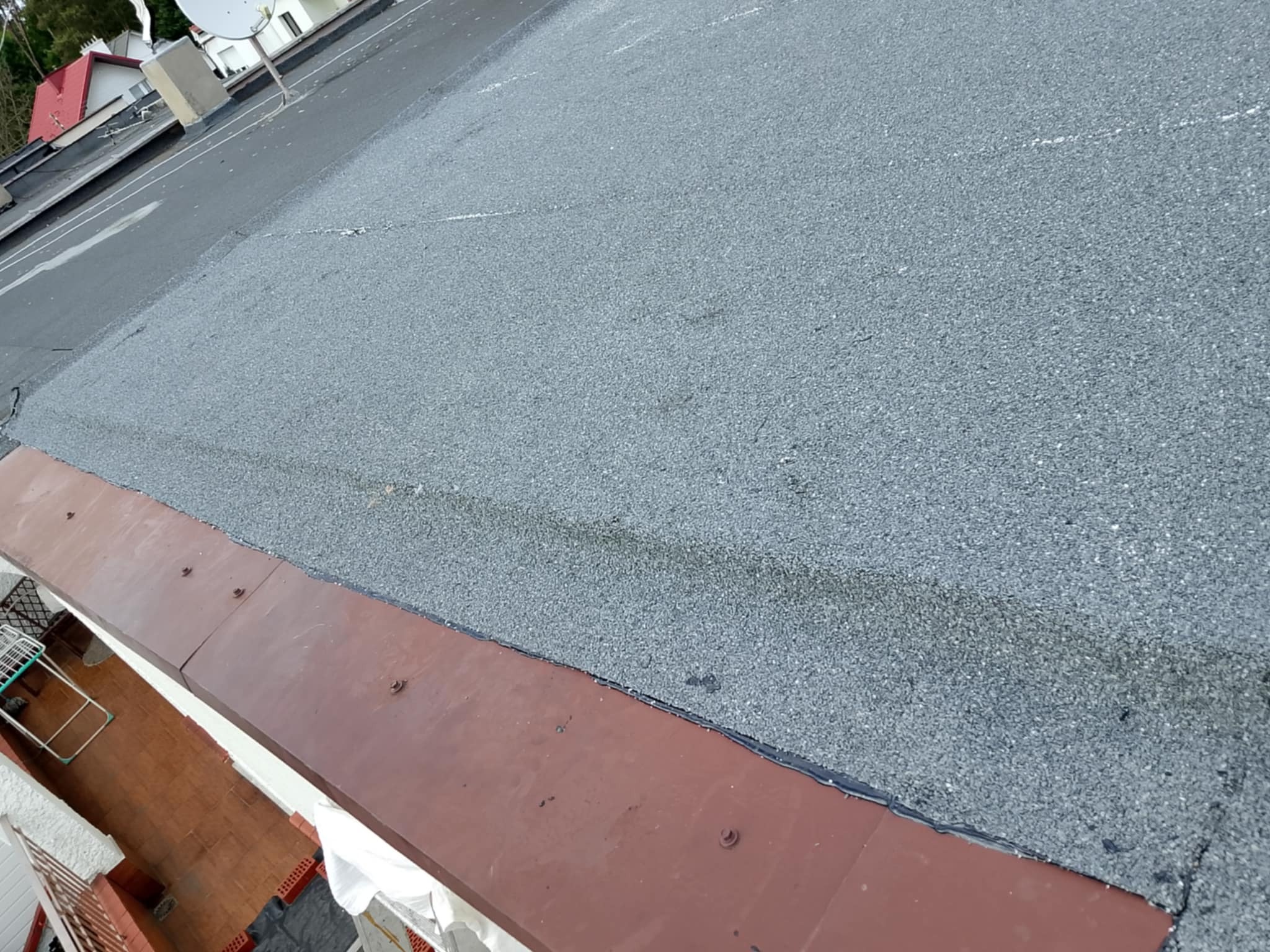 Szary dach pokryty papą z widocznym brązowym obrzeżem i fragmentem balkonu. Ujęcie z góry, prezentujące wykończenie dachu płaskiego.