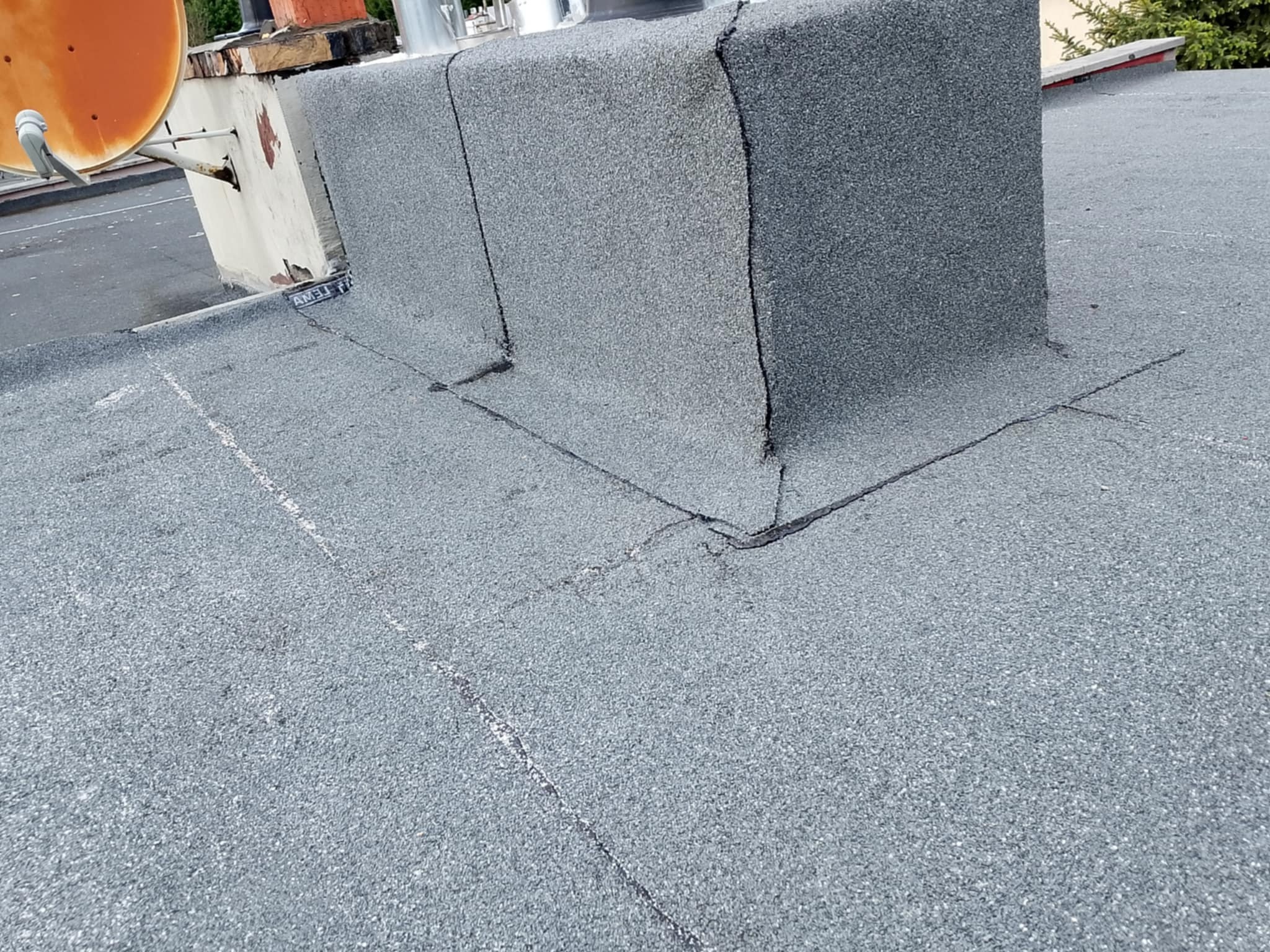 Szary dach pokryty papą z kominem i anteną satelitarną. Widoczne łączenia papy i detale wykończenia komina. Lekki kąt ujęcia.