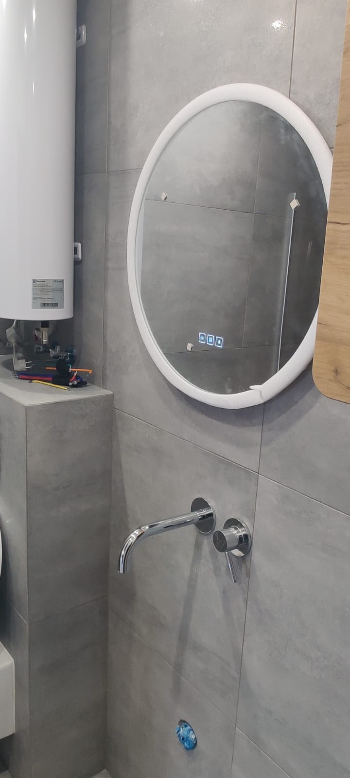 Wykończona łazienka: szare płytki, okrągłe lustro z podświetleniem, bateria ścienna i bojler. Widoczne narzędzia sugerują prace wykończeniowe.