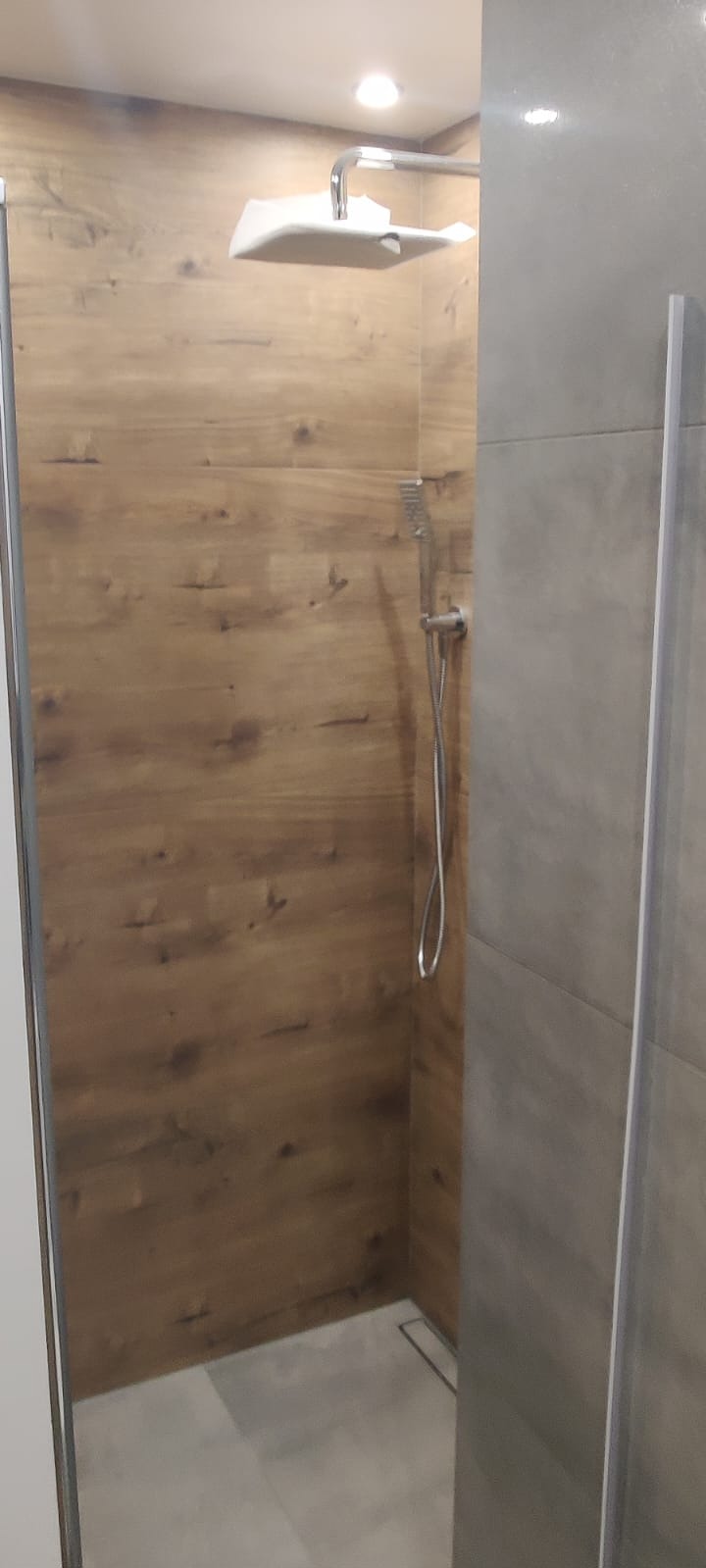 Nowoczesna kabina prysznicowa z drewnopodobną okładziną ścienną i szarą glazurą. Widoczny odpływ liniowy i deszczownica.