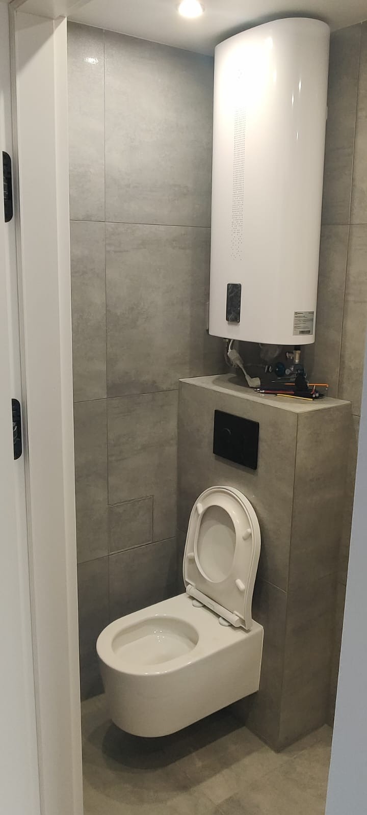 Szara łazienka z podwieszaną toaletą i białym bojlerem. Na podeście bojlera leżą narzędzia. Widoczne szare płytki i białe drzwi.