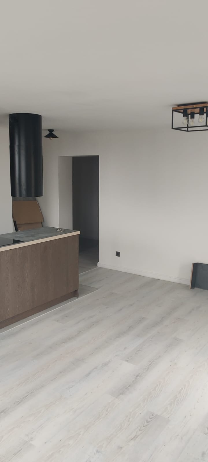 Jasne panele podłogowe w nowym wnętrzu z ciemnymi akcentami: okap kuchenny i lampa sufitowa. Widoczny fragment zabudowy kuchennej i drzwi do innego pomieszczenia.