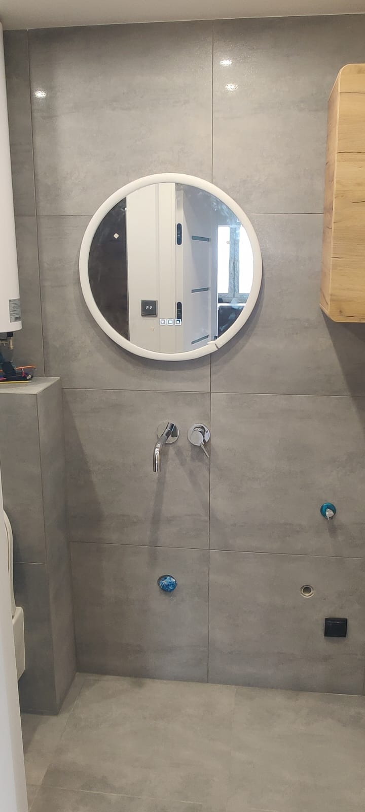 Szare płytki w łazience z okrągłym lustrem i wystającymi kranami. Widoczne niebieskie zaślepki i otwory na armaturę, sugerujące etap prac wykończeniowych.