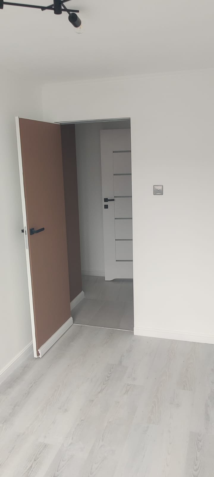 Nowoczesne wnętrze z drewnianą podłogą i dwiema parami drzwi: brązowe otwarte i białe zamknięte z poziomymi frezami. Minimalistyczny design i oświetlenie sufitowe.