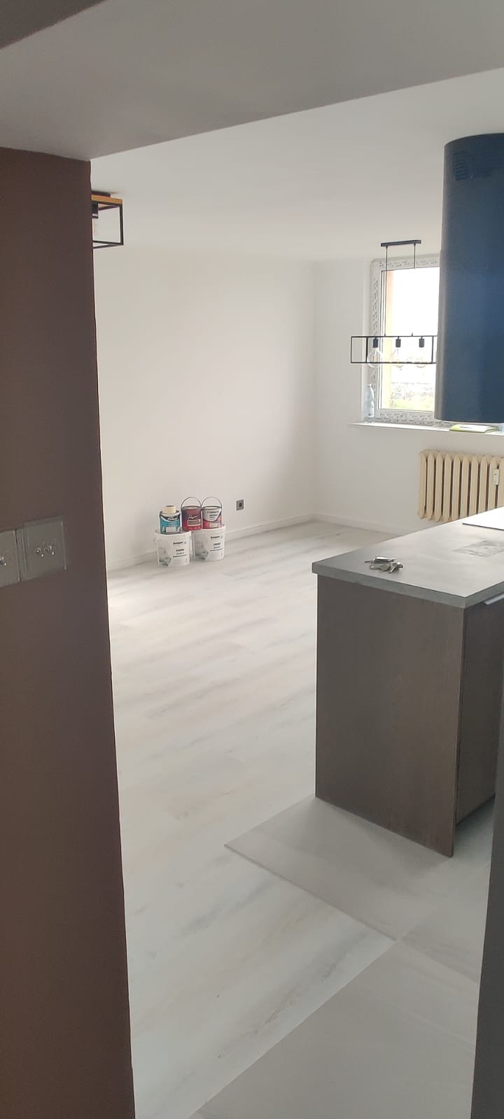 Jasne, odmalowane wnętrze z podłogą imitującą drewno, widoczne puszki z farbą. Widok z perspektywy wejścia do pomieszczenia.