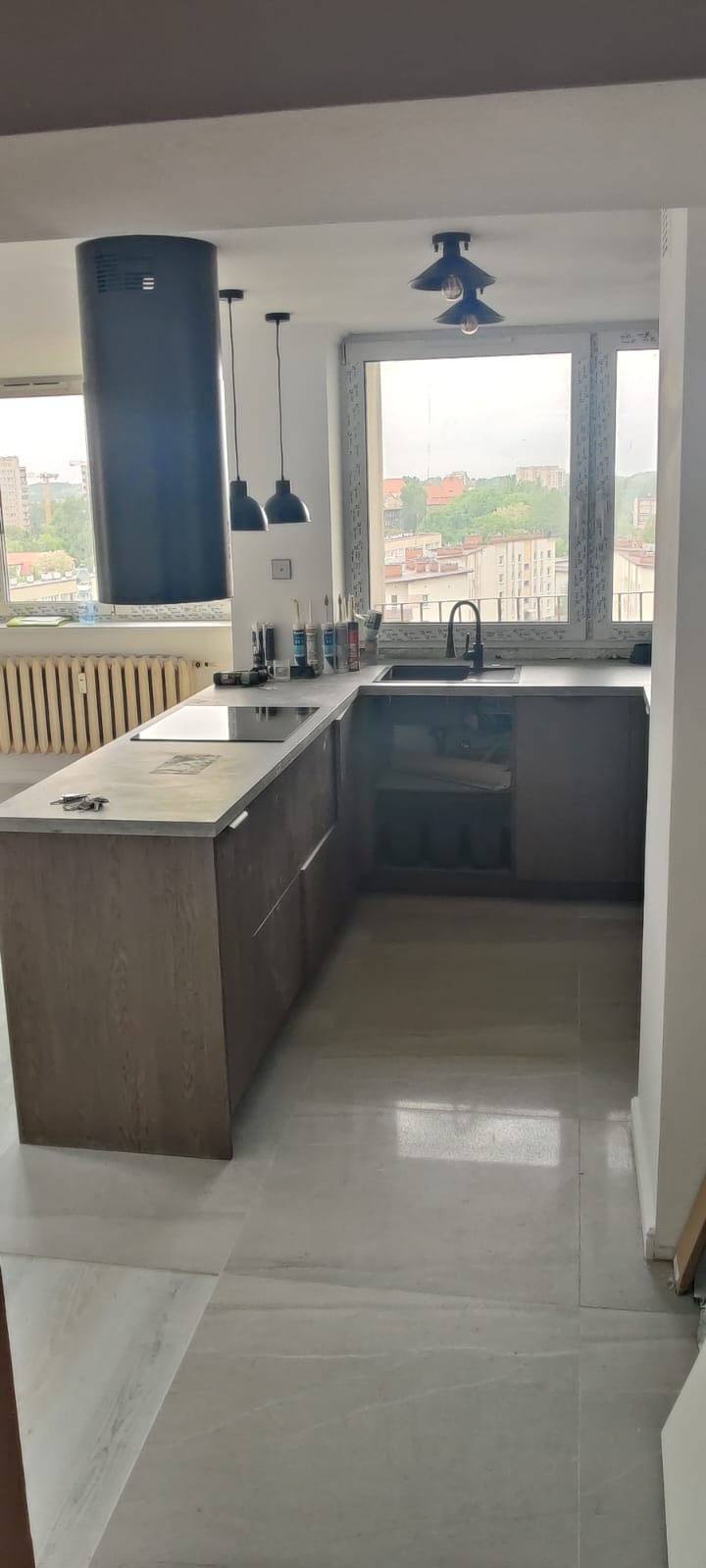 Nowoczesna kuchnia z wyspą i widokiem na miasto. Szare płytki podłogowe i drewniane szafki. Nad wyspą wiszą czarne lampy, a w tle widać zabudowę miejską.