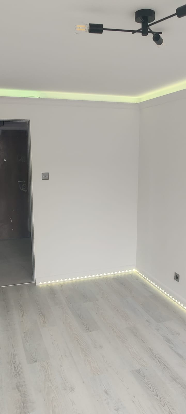 Białe wnętrze po remoncie z panelami, listwami LED na suficie i podłodze, minimalistyczną lampą sufitową i widocznym fragmentem surowej ściany w przejściu.