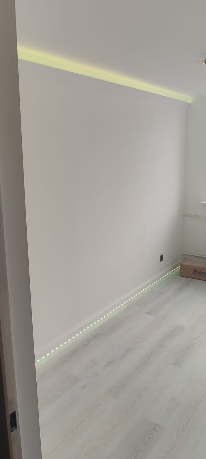 Białe wnętrze z listwami LED: sufitowa i przypodłogowa, oświetlające ścianę. Widoczne panele podłogowe i fragment drzwi po lewej stronie.