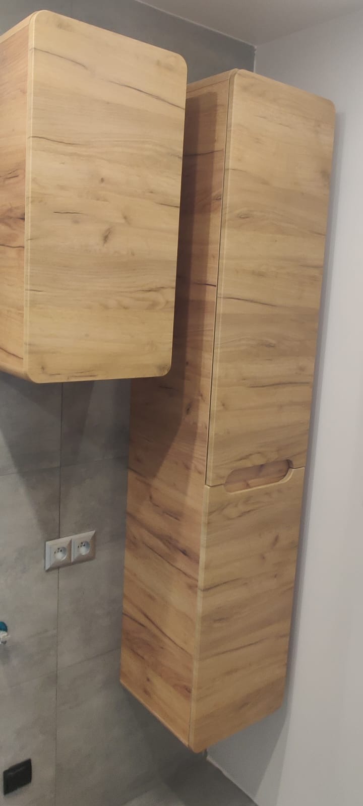 Wykończona łazienka: drewnopodobne szafki wiszące z frezowanymi uchwytami, szara płytka ścienna i gniazdko elektryczne. Ujęcie z dołu.