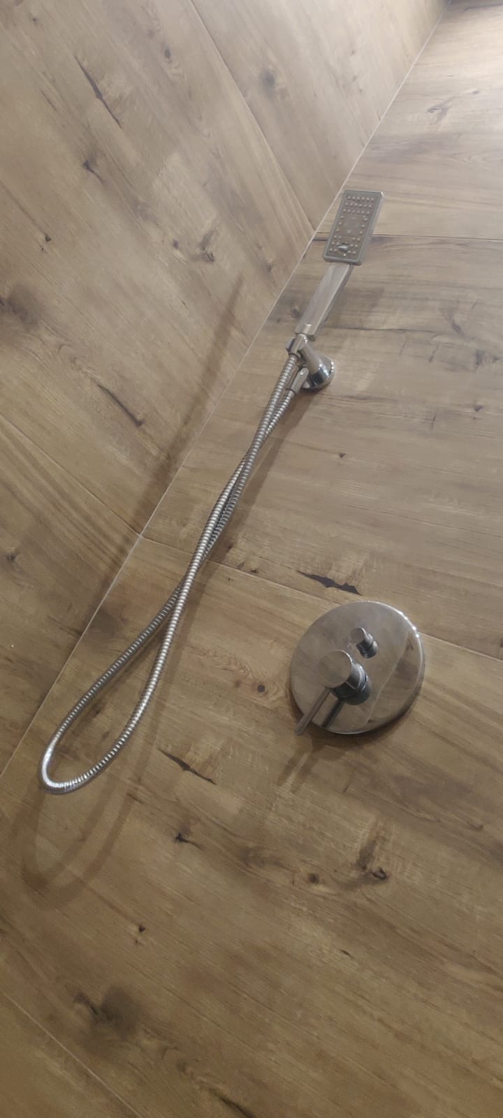 Nowoczesna bateria prysznicowa z deszczownicą i okrągłym panelem sterującym, zamontowana na ścianie wyłożonej płytkami imitującymi drewno.