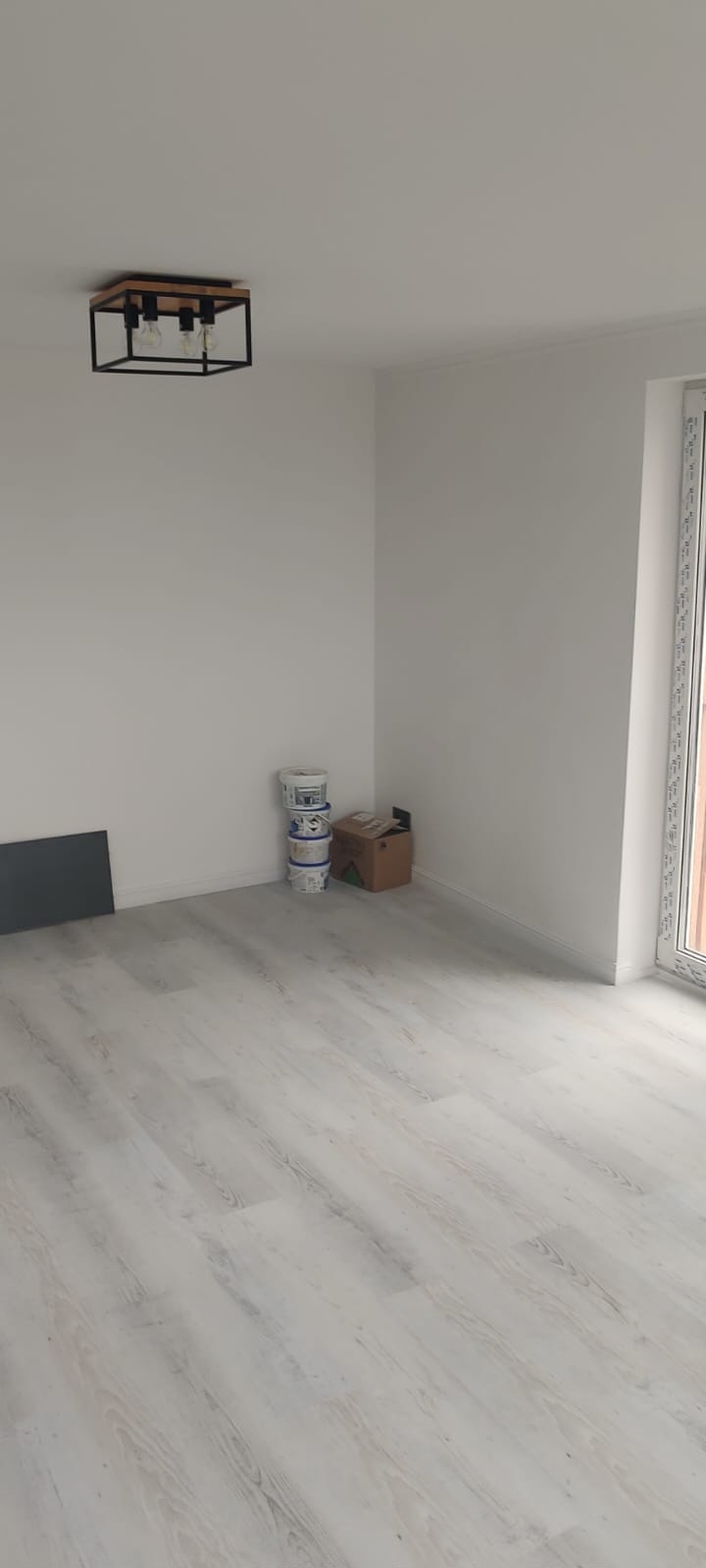 Jasne panele podłogowe w pustym pokoju po remoncie, widoczne puszki z farbą i nowoczesna lampa sufitowa. Świeżo pomalowane ściany na biało i okno z białymi ramami.