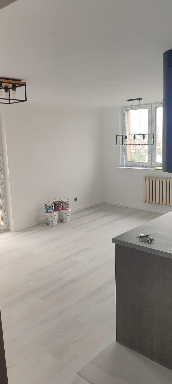 Jasne panele podłogowe w świeżo pomalowanym na biało pokoju z oknem. Widoczne puszki z farbą, grzejnik i nowoczesne lampy.