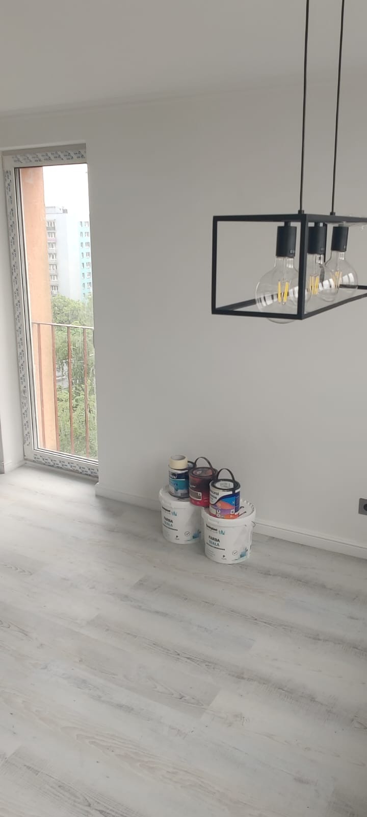 Jasne panele podłogowe i świeżo pomalowane białe ściany w pokoju z oknem i nowoczesną lampą. Widoczne puszki z farbą.