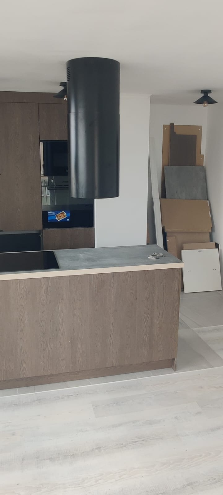 Nowoczesna kuchnia w trakcie montażu: wyspa z szarym blatem, czarny okrągły okap i drewniane fronty. W tle płyty do zabudowy.