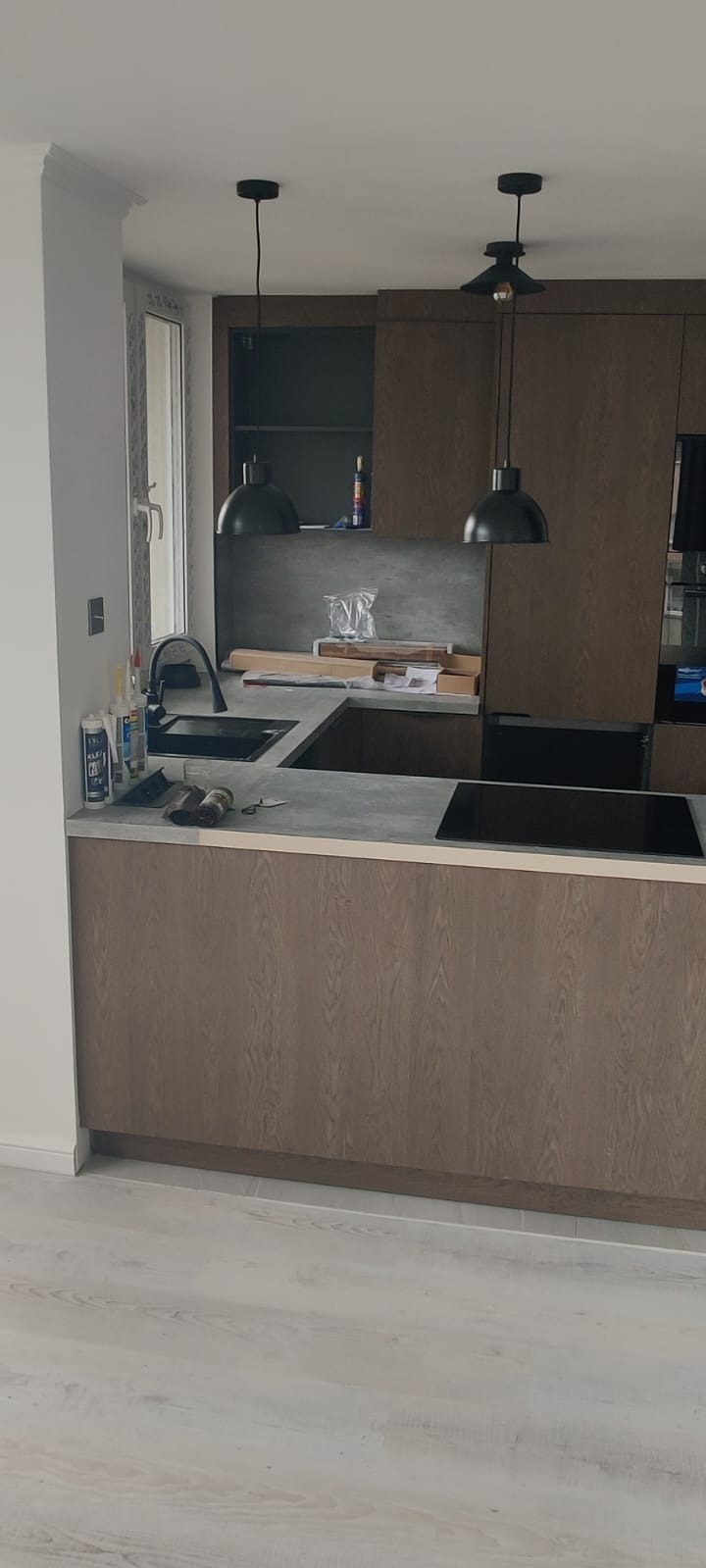 Nowoczesna kuchnia z drewnianymi szafkami, czarnym zlewem i lampami wiszącymi. Na blacie widoczne narzędzia i akcesoria montażowe. Jasne podłogi.