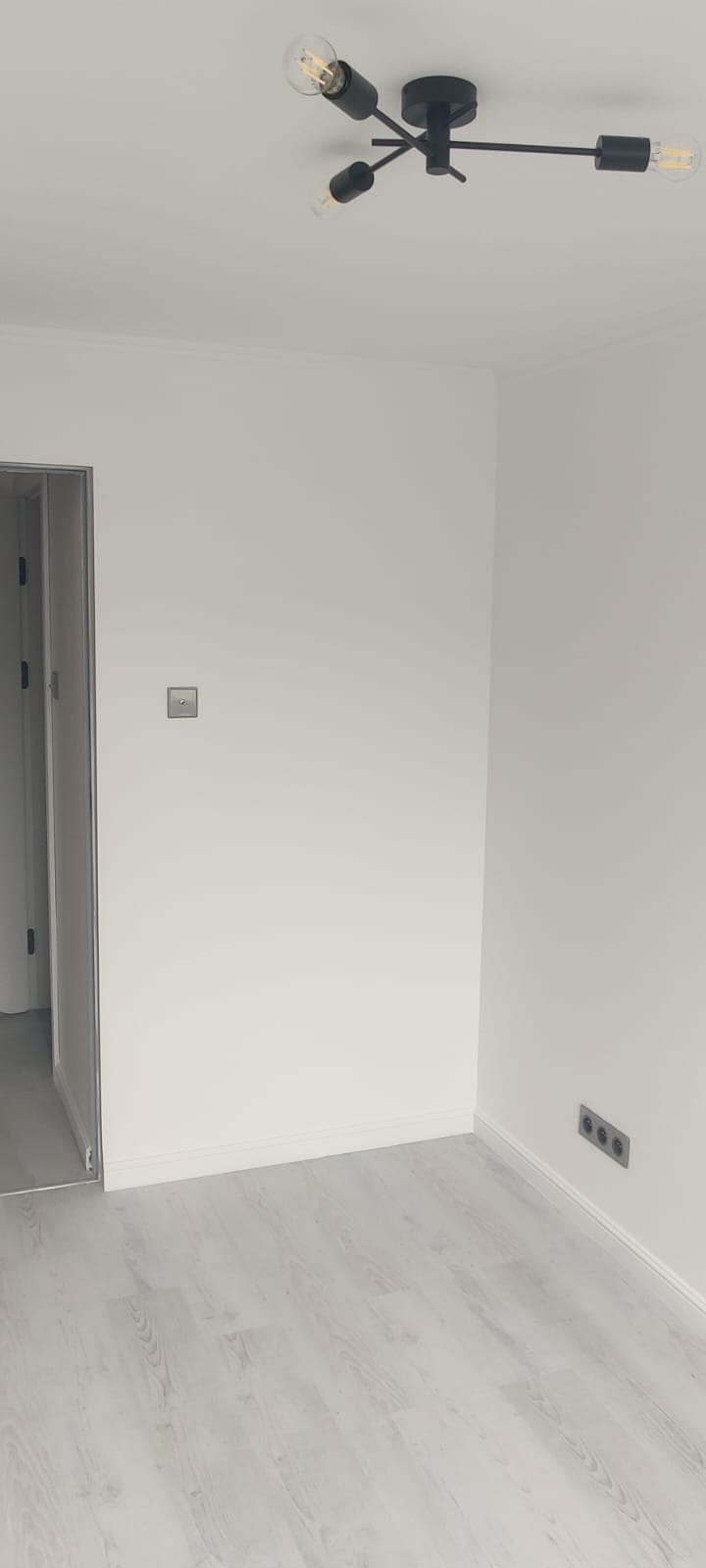 Minimalistyczne wnętrze: białe ściany, jasna podłoga, czarna lampa sufitowa z żarówkami Edisona i szara listwa przypodłogowa. Widoczne gniazdka elektryczne.