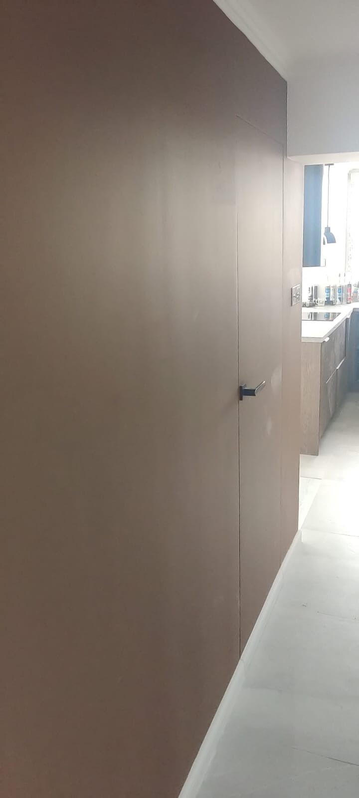 Ukryte drzwi w brązowej ścianie, minimalistyczny design. Czarny, prosty uchwyt. W tle widoczna kuchnia w jasnych barwach.