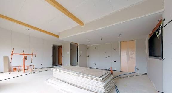 Wnętrze w trakcie prac wykończeniowych: ściany obłożone płytami gips-karton, widoczny podnośnik i stos płyt. Surowy stan, przygotowanie do dalszych prac.