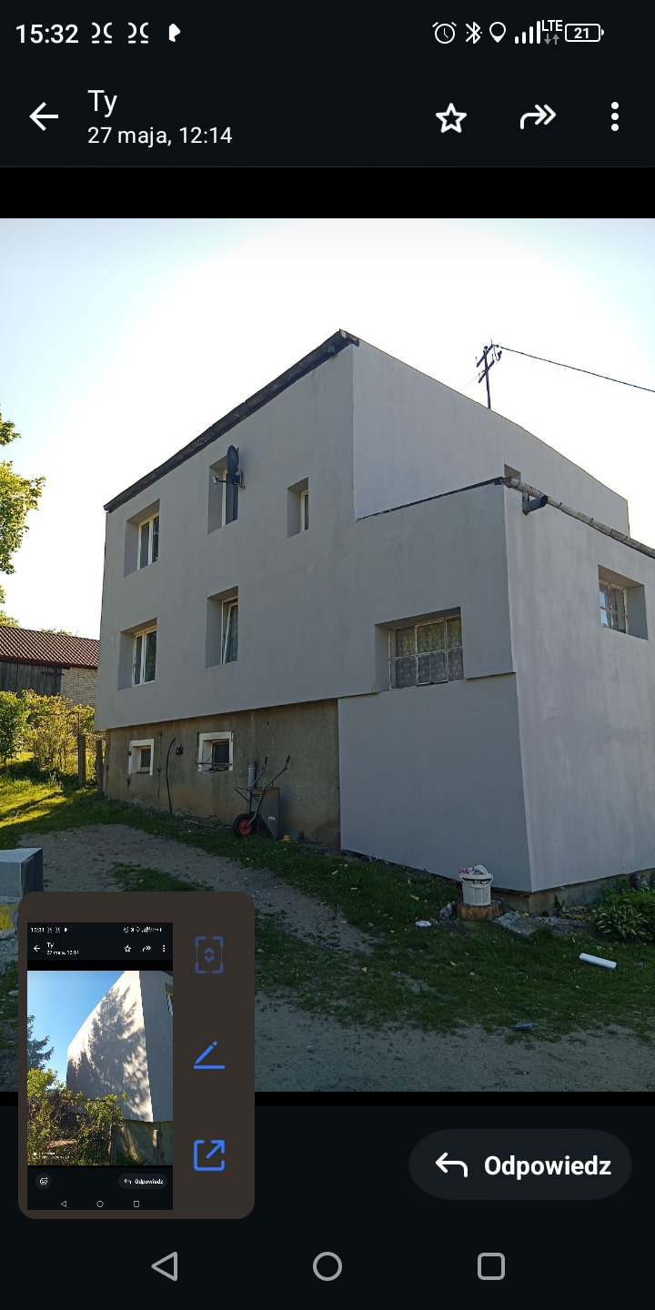 Dwupiętrowy dom po szpachlowaniu i malowaniu elewacji na szaro, z widocznymi oknami i satelitą. W tle zieleń i fragment budynku gospodarczego.