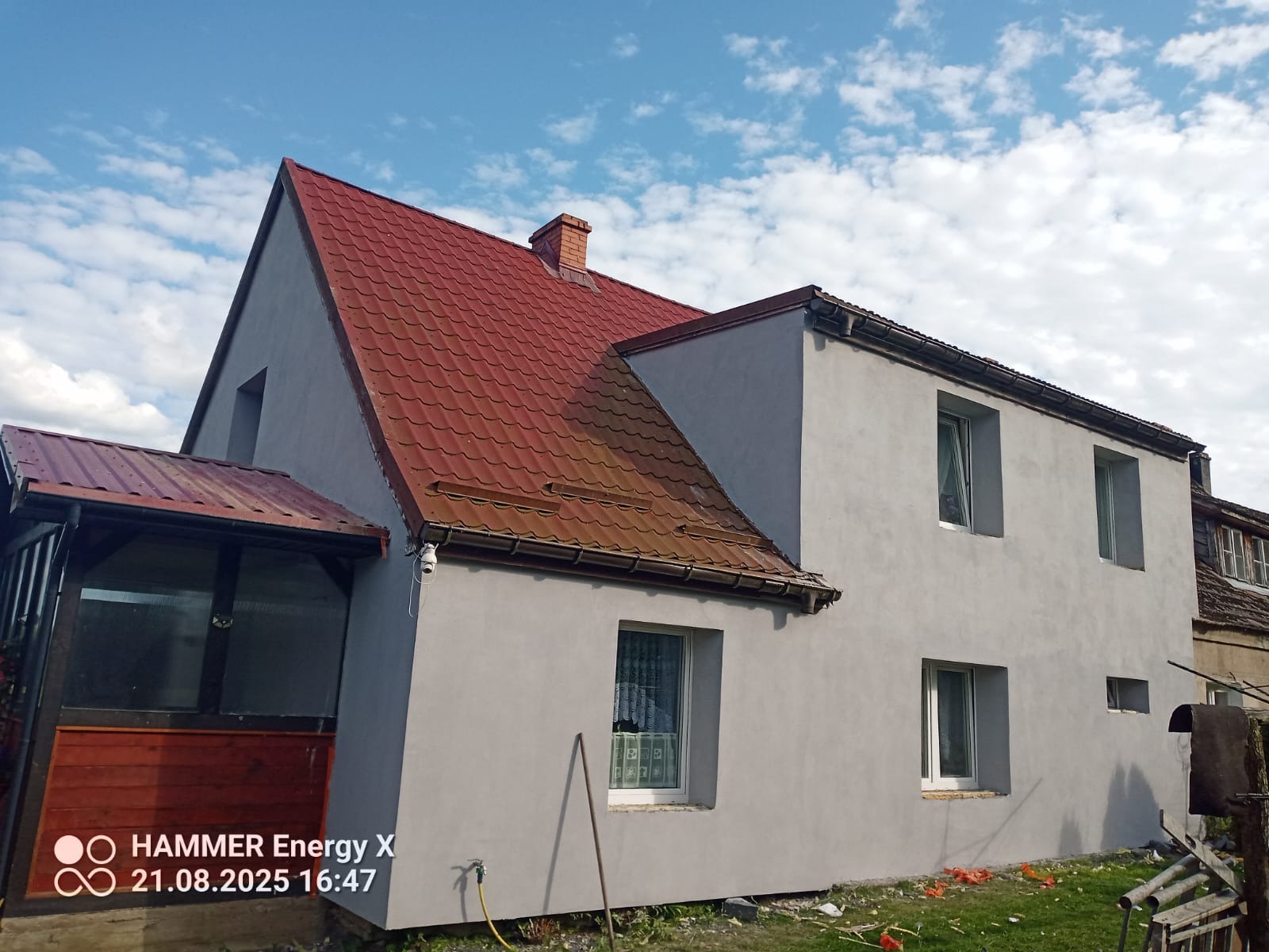 Odnowiony dom z czerwoną dachówką i szarą elewacją, widoczny z zewnątrz w słoneczny dzień. Nowe okna, świeżo otynkowana ściana i fragment werandy.