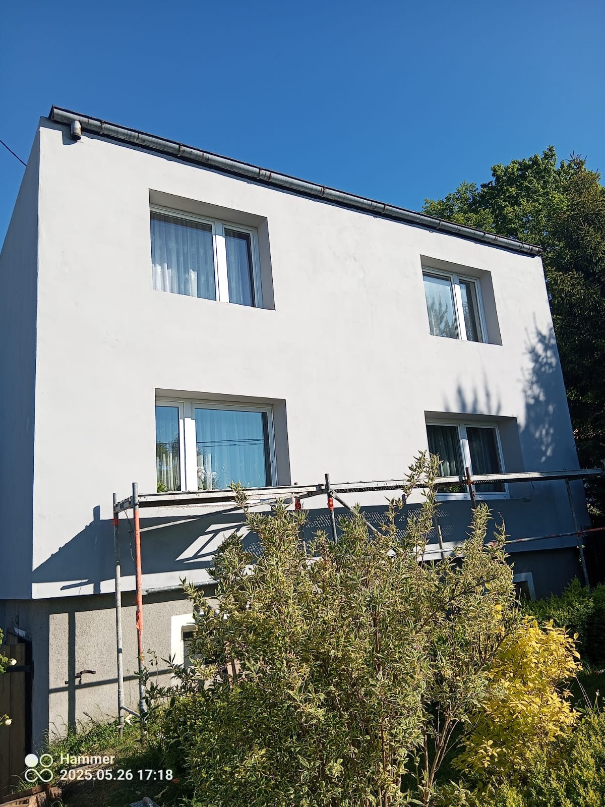 Dwukondygnacyjny budynek po szpachlowaniu, z oknami i rusztowaniem, widok z dołu, błękitne niebo, zieleń drzew, krzew na pierwszym planie.