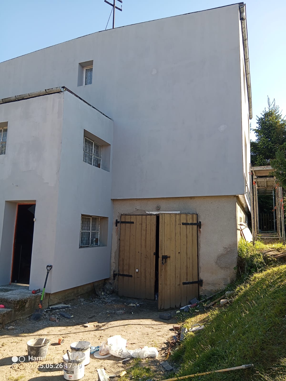 Elewacja budynku po malowaniu na szaro, widoczne drewniane drzwi do garażu, narzędzia malarskie. Ujęcie z dołu, w tle zieleń i rusztowanie.