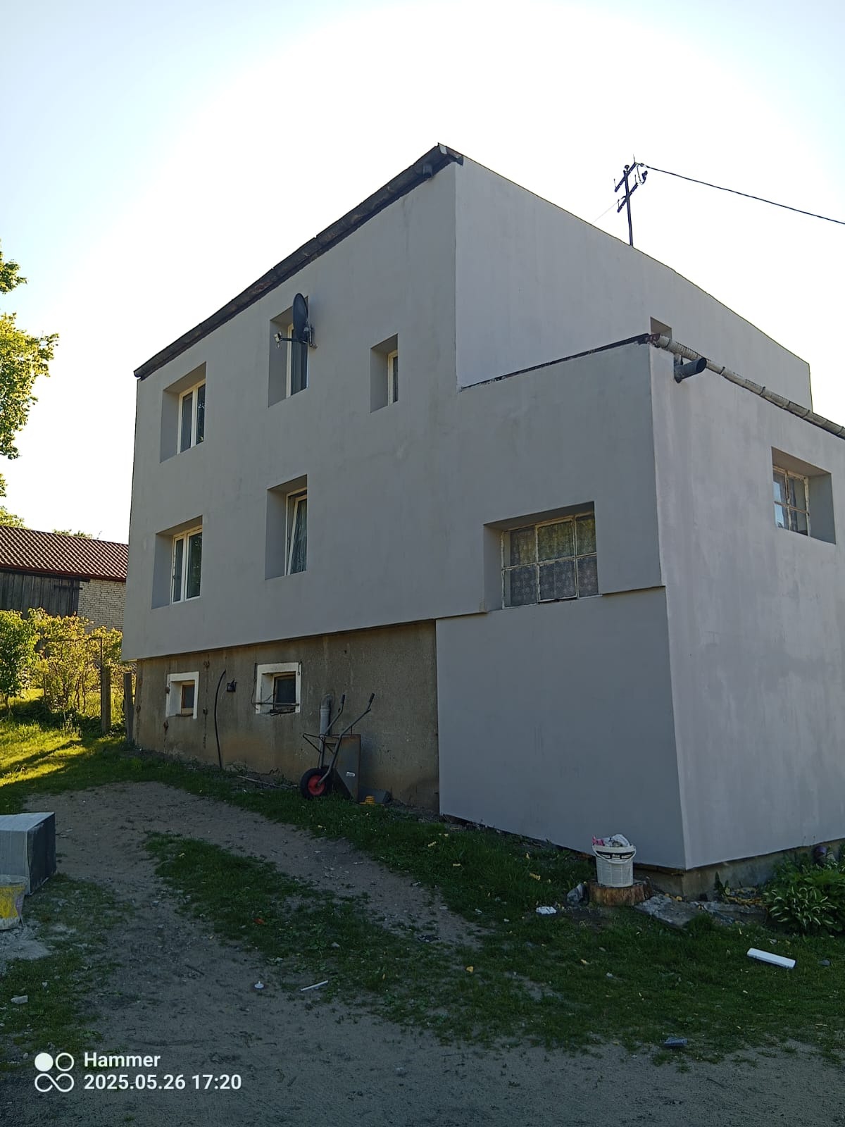 Otynkowany, dwukondygnacyjny budynek z szarą elewacją w słoneczny dzień. Widoczna antena satelitarna i rynna. Na trawniku wiadro i taczka.