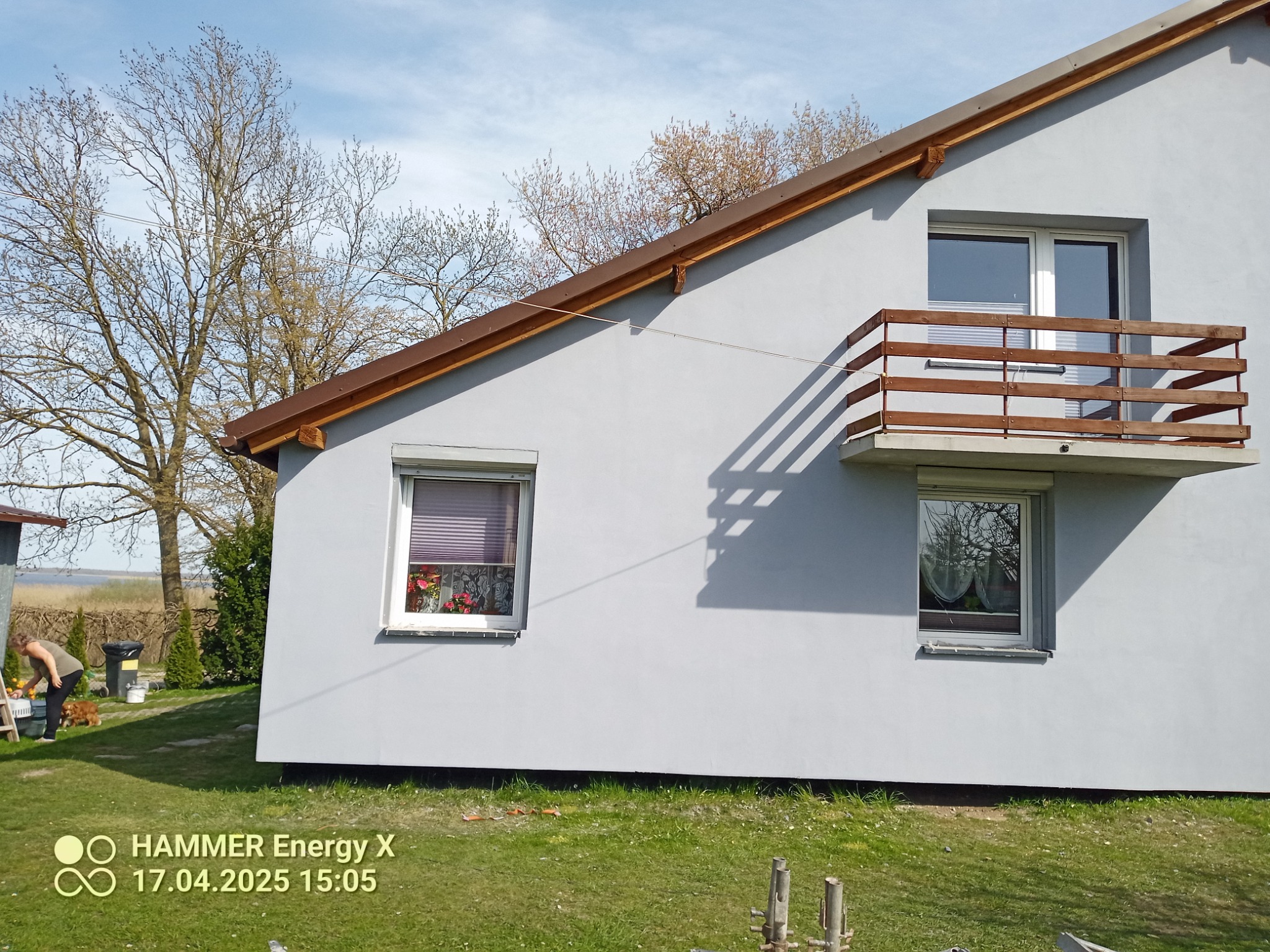 Szara elewacja domu z drewnianą balustradą balkonu w Dębnicy Kaszubskiej. Widoczne okna z roletami i kwiatami, oraz osoba w ogrodzie.