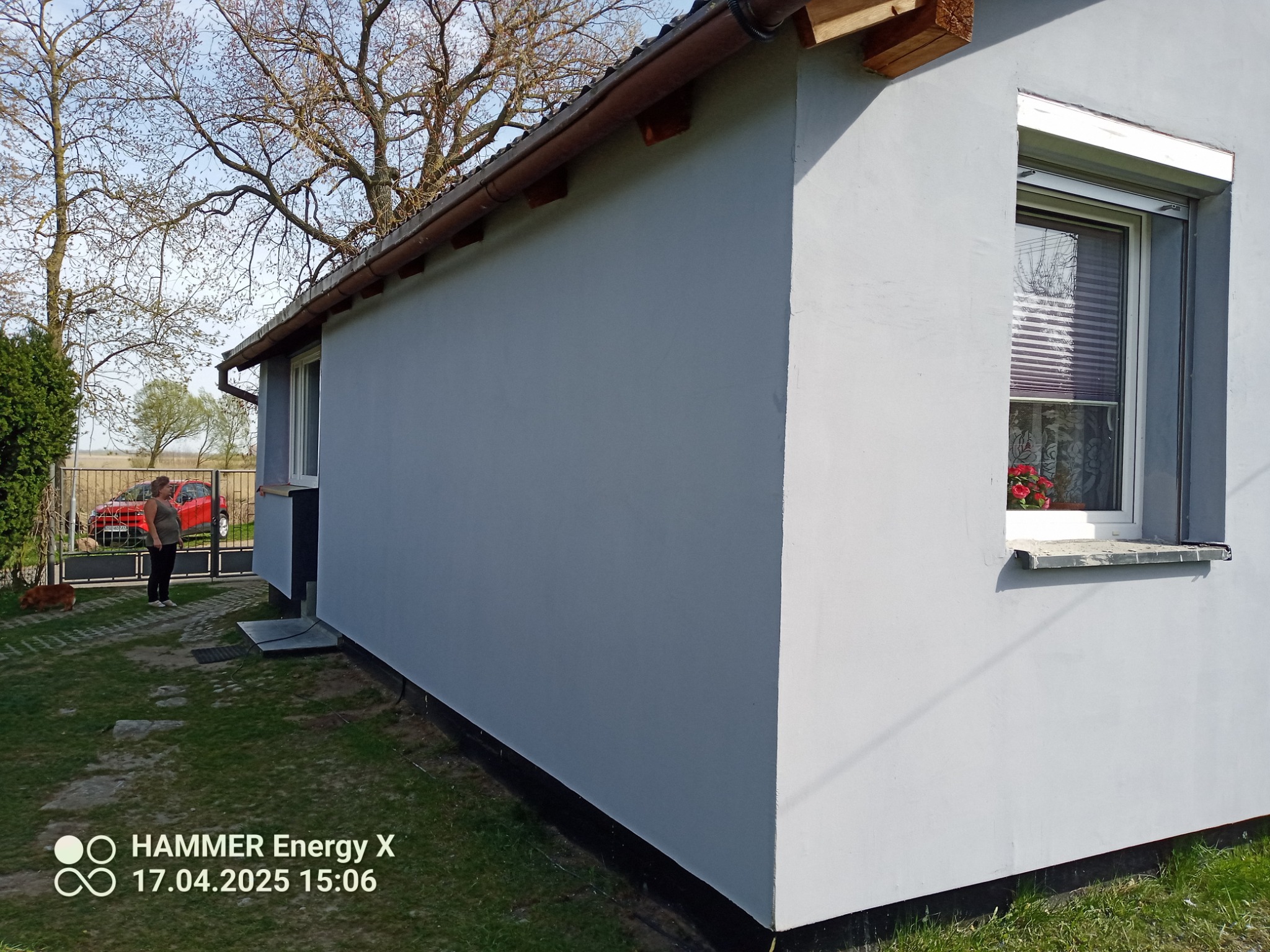 Ściana budynku po szpachlowaniu i malowaniu na szaro, z oknem z roletą i kwiatami. Widoczna osoba i czerwony samochód w tle, zieleń trawy.