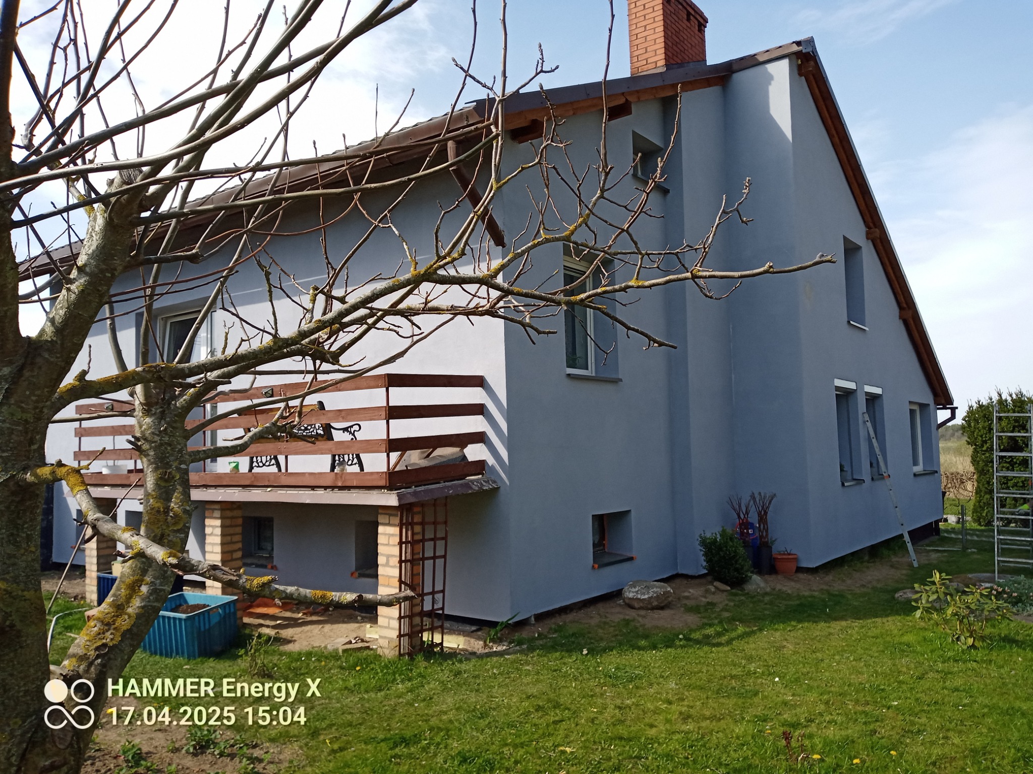 Szary dom jednorodzinny z ceglanym kominem i drewnianym balkonem, widziany z perspektywy ogrodu, z gałęziami drzewa na pierwszym planie. Widoczne drabiny i donice.