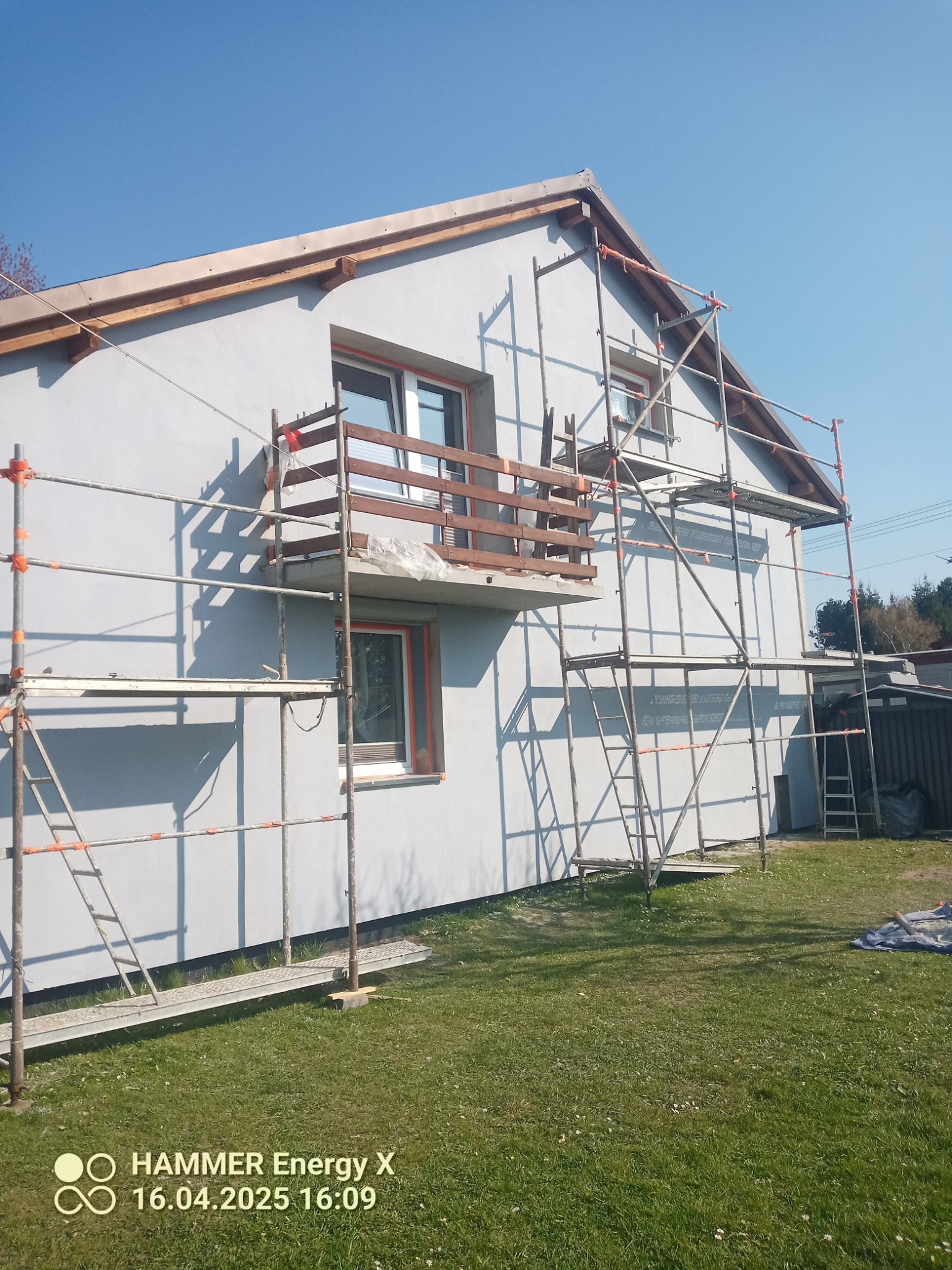 Dom jednorodzinny w trakcie prac elewacyjnych, otoczony rusztowaniem. Widoczny balkon z drewnianą balustradą i świeżo pomalowana fasada na tle błękitnego nieba.